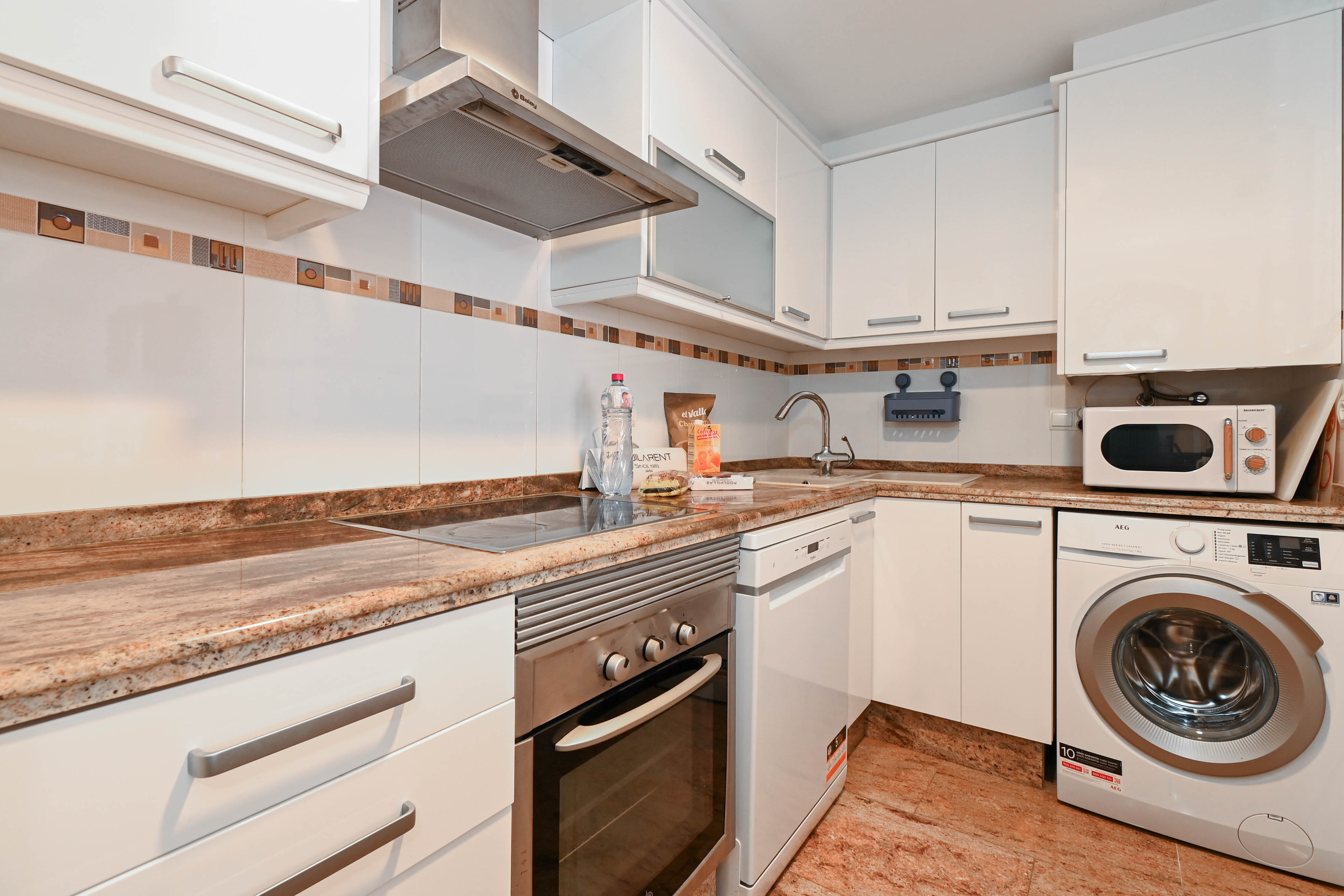 Functionele keuken met witte kasten, granieten werkbladen en inbouwapparatuur waaronder een wasmachine, vaatwasser en roestvrijstalen oven.