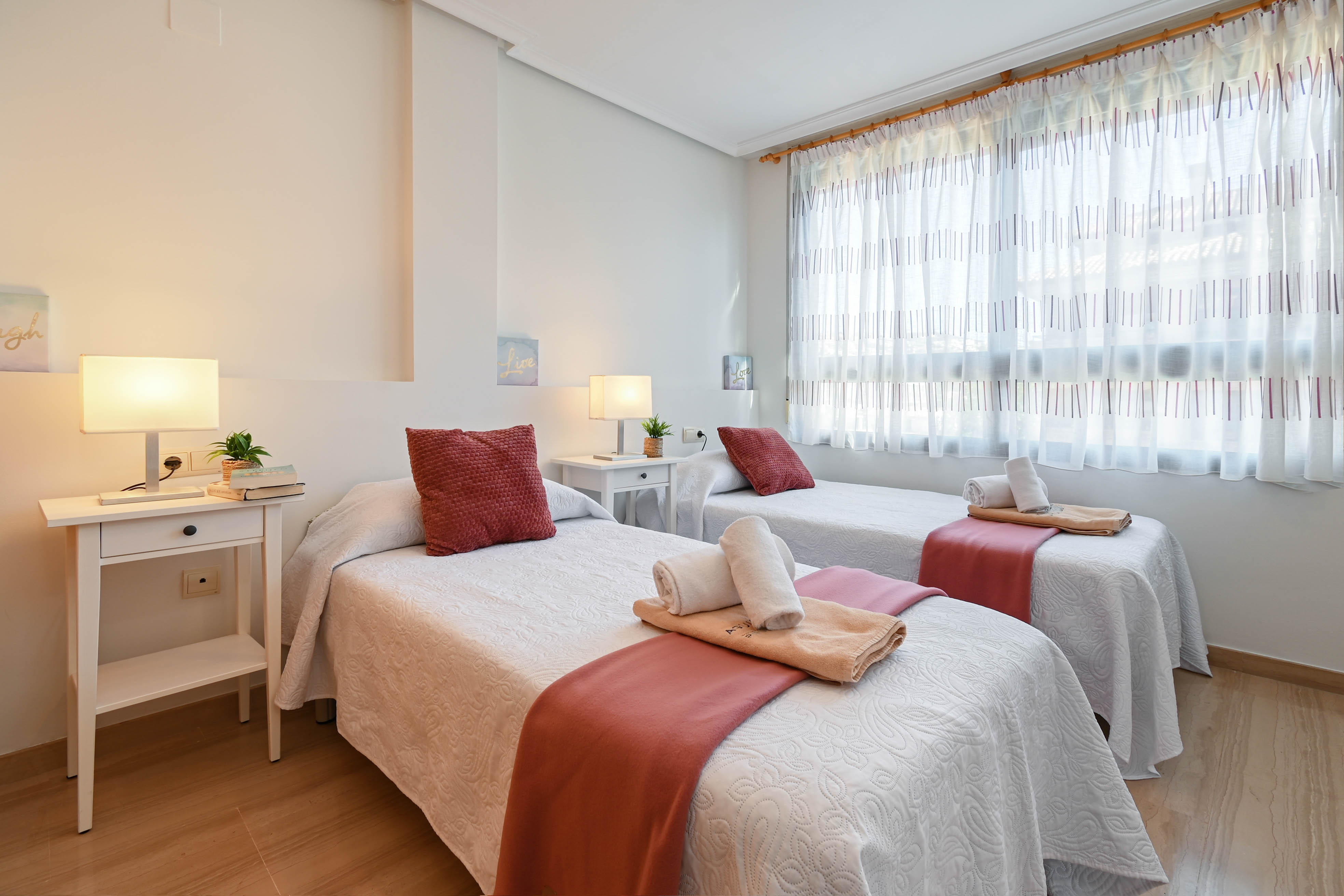 Lichte slaapkamer met twee eenpersoonsbedden, witte nachtkastjes en een lichte houten vloer. Een groot raam met transparante gordijnen biedt volop natuurlijk licht.