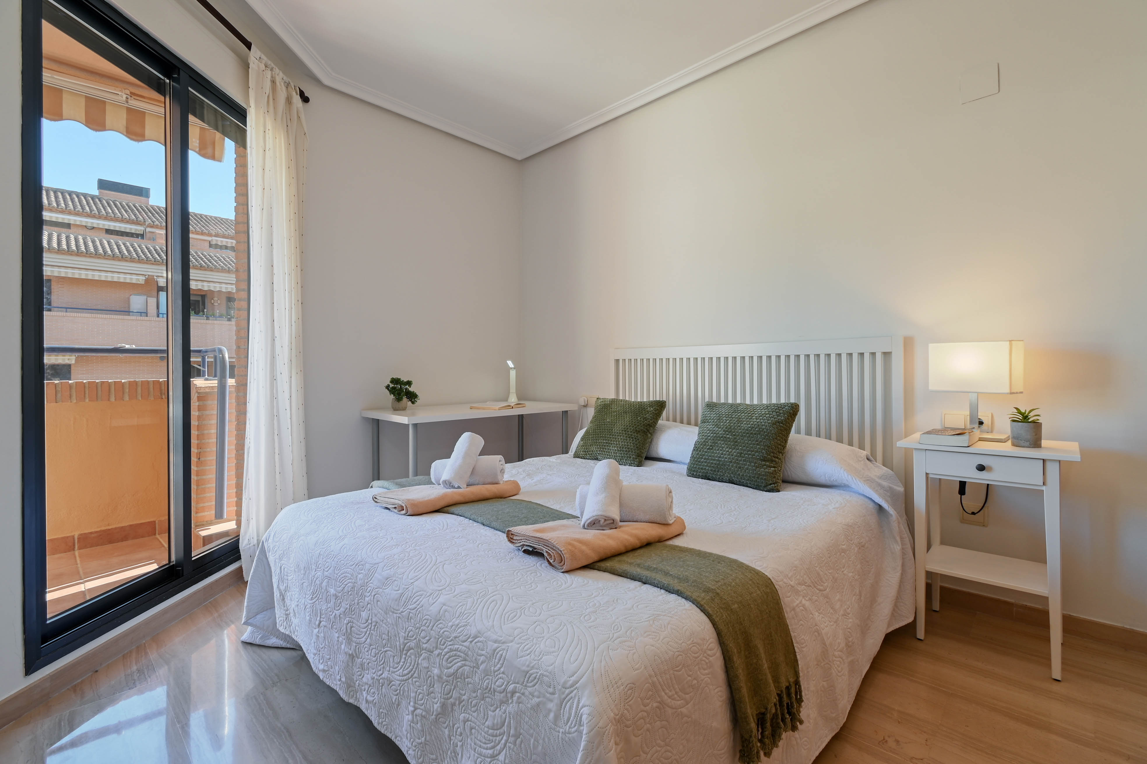 Lichte slaapkamer met tweepersoonsbed, wit lattenhoofdbord, bureau en directe toegang tot een privébalkon via grote glazen schuifdeuren.