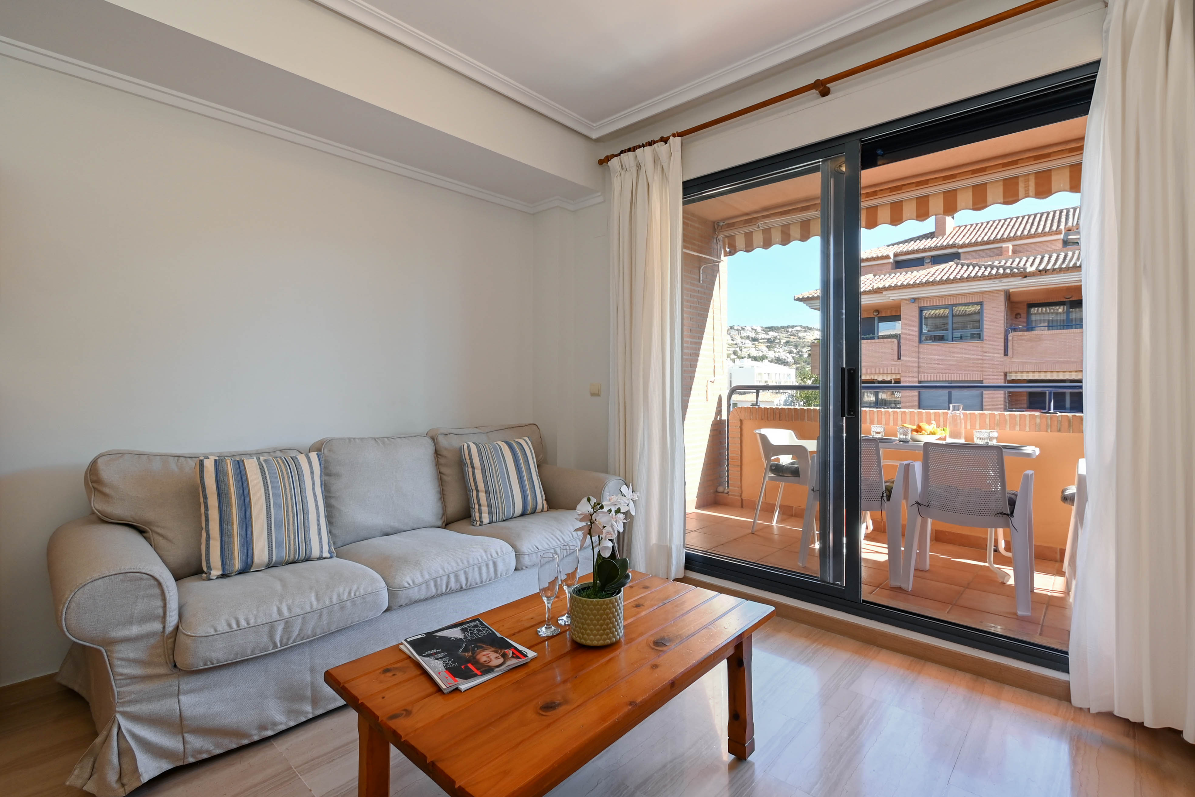 Lichte woonkamer met een grijze bank en houten salontafel, verbonden via een schuifpui met een gemeubileerd balkon met uitzicht op de omgeving.