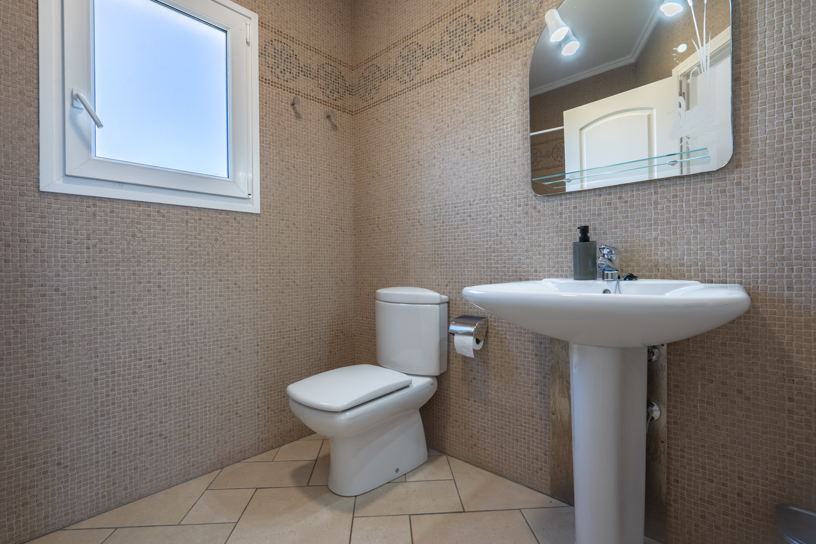 Badkamer met beige mozaïektegels, een wastafel op zuil en een wit PVC-raam voor natuurlijke lichtinval.