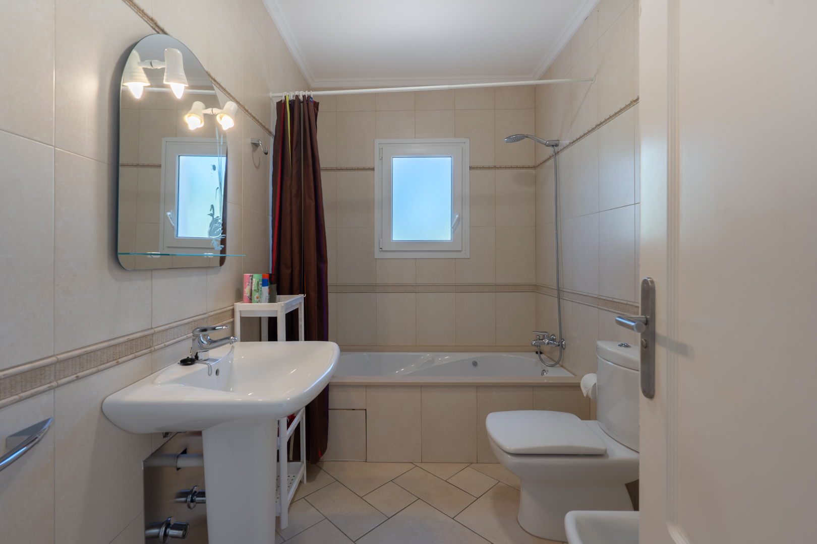Badkamer met beige wandtegels, een ingebouwd ligbad, een wastafel op zuil en een boogspiegel met geïntegreerde verlichting naast een PVC-raam.