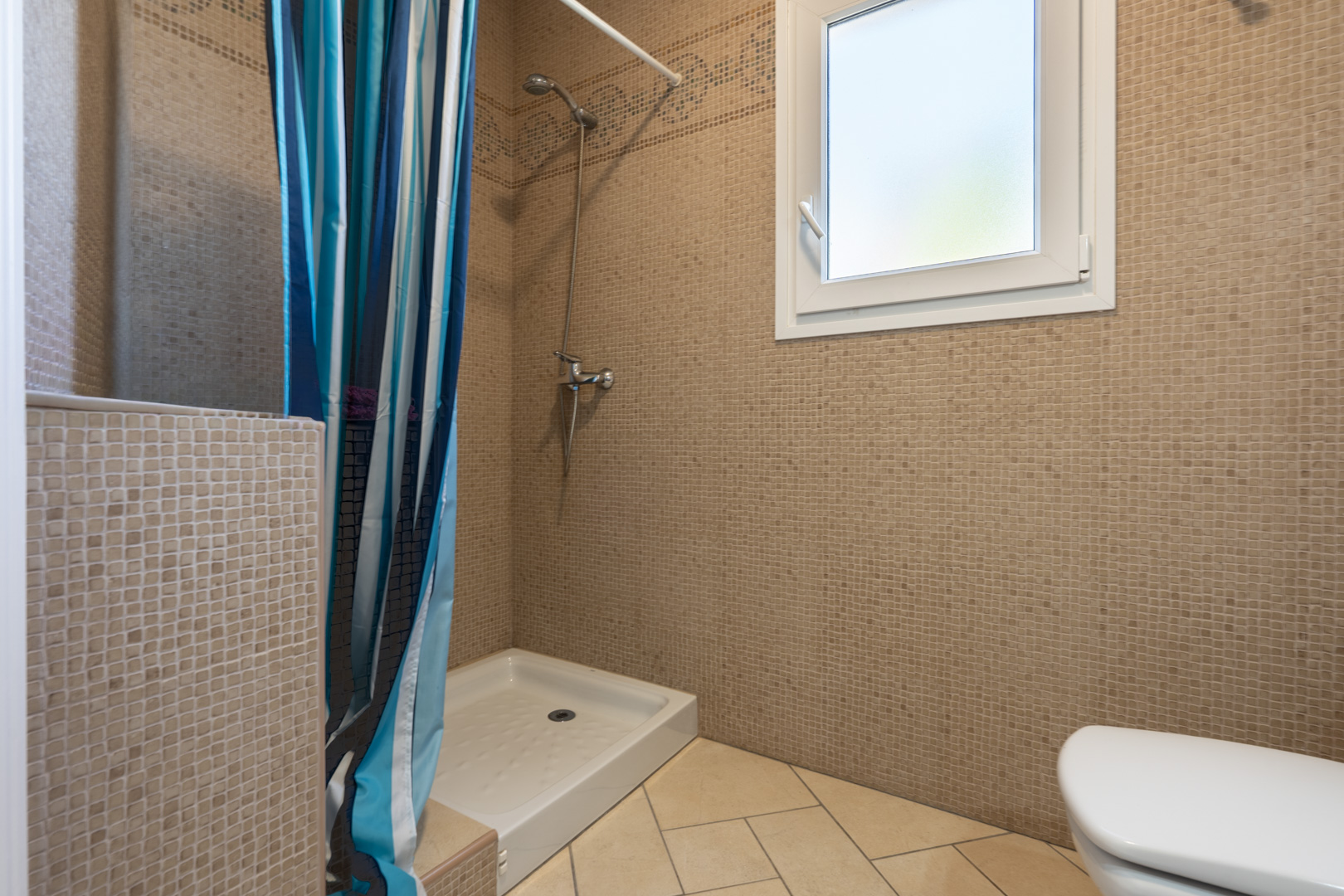 Badkamer met bruine mozaïektegels, een witte douchebak met gestreept gordijn en een wit PVC-raam dat zorgt voor natuurlijke lichtinval.