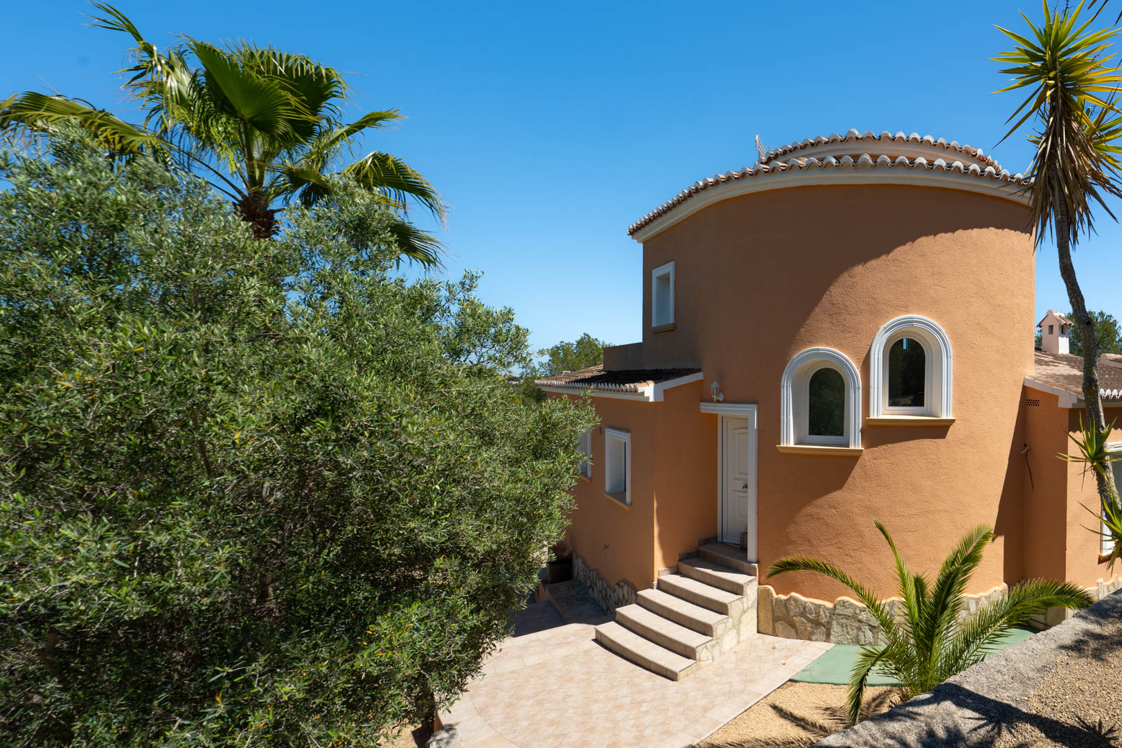 Exterieur van de villa met een terracotta gevel, een ronde torenstructuur met boogvensters en stenen treden die naar de voordeur leiden.