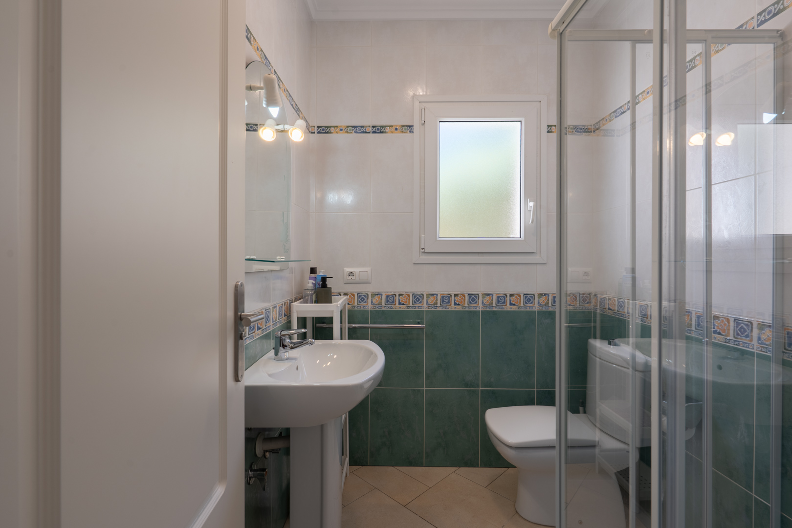 Functionele badkamer met groen-witte betegeling, een kolomwastafel, glazen douchecabine en een PVC-raam met matglas.