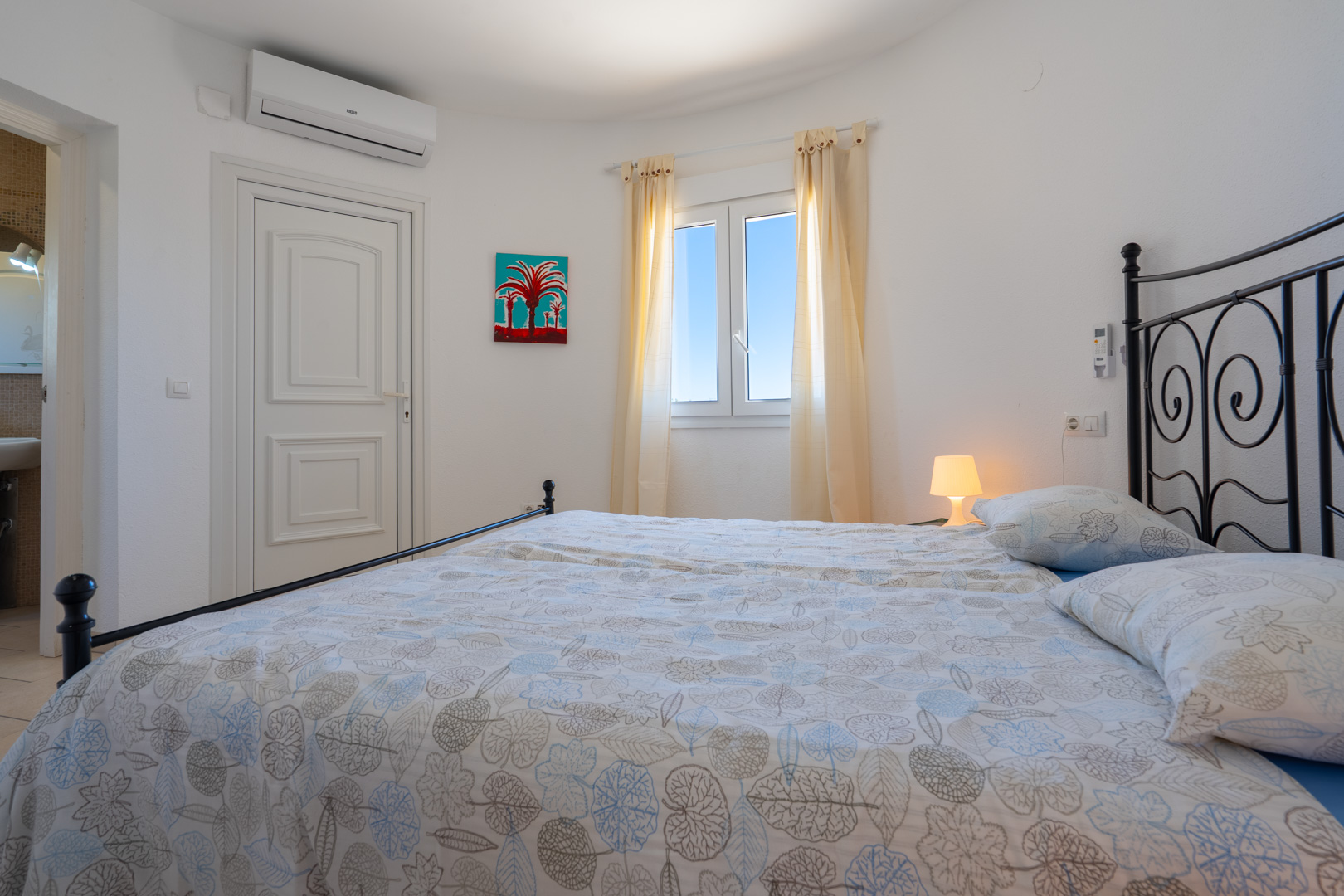 Lichte slaapkamer met een zwart smeedijzeren bedframe, airconditioning, PVC-raam en directe toegang tot de aangrenzende badkamer.