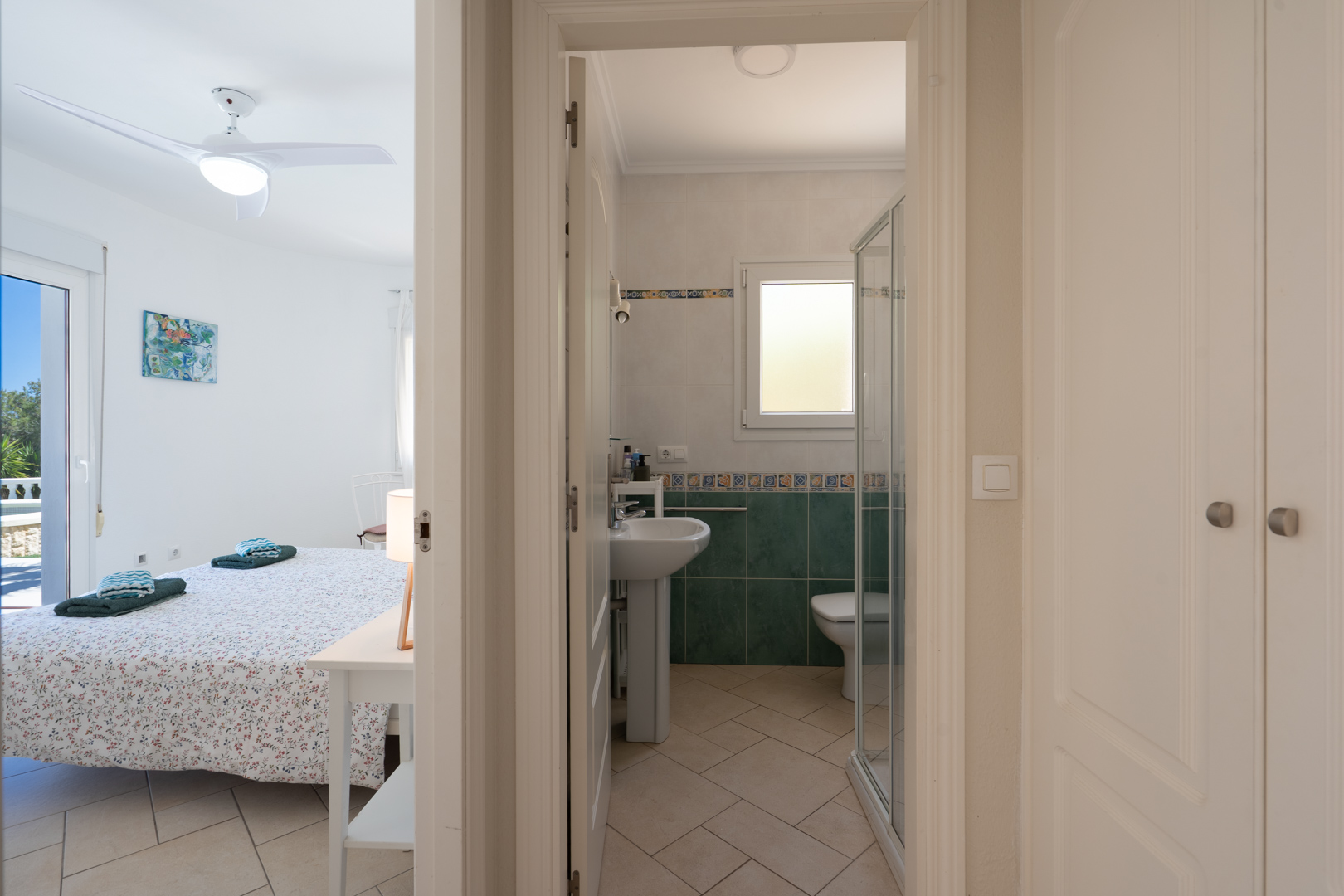 Lichte slaapkamer met plafondventilator en directe toegang tot buiten, gelegen naast een badkamer met douche en ingebouwde kasten in de gang.
