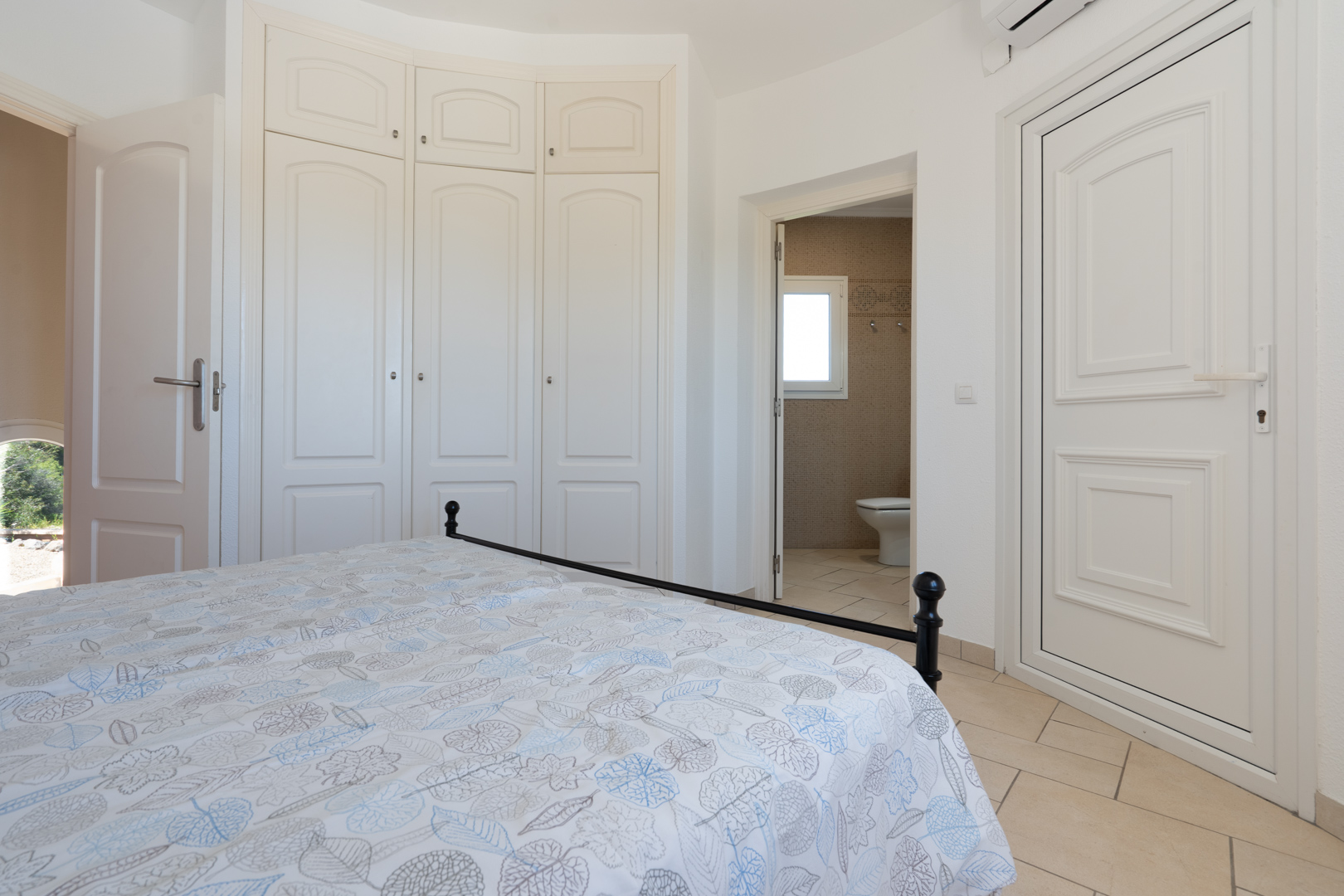 Lichte slaapkamer met witte inbouwkasten, keramische tegelvloer en directe toegang tot een aangrenzende badkamer met mozaïektegels.