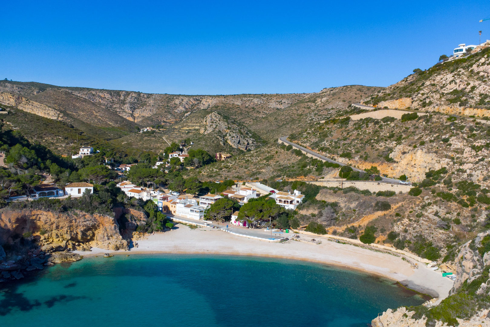 Luchtfoto van Cala Granadella in Jávea, met kristalhelder turquoise water, een kiezelstrand en witte woningen verscholen tussen de mediterrane rotsen.