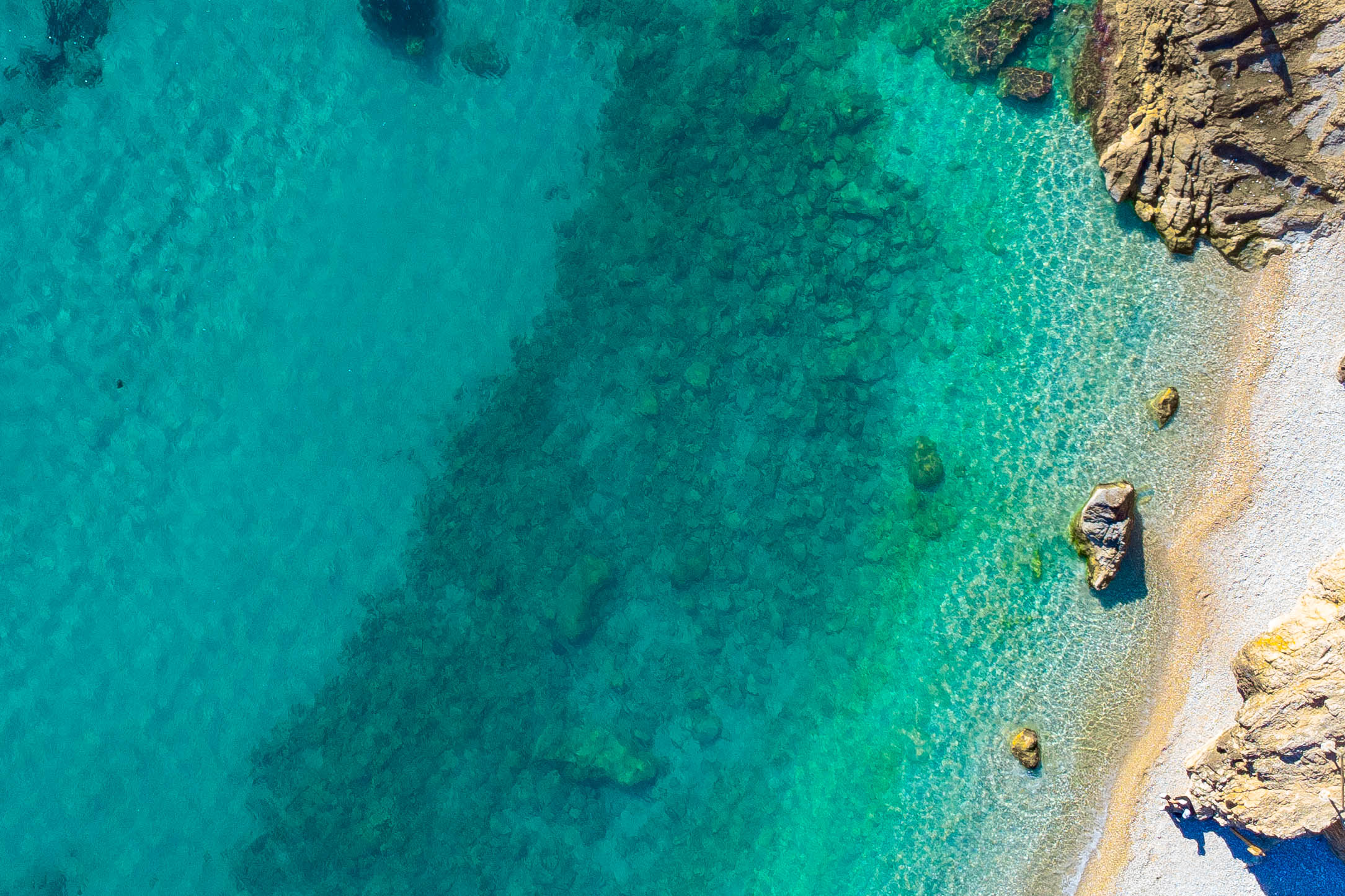 Luchtfoto van de kustlijn van Jávea met kristalhelder turquoise water, een zichtbare rotsachtige zeebodem en een natuurlijk kiezelstrand.