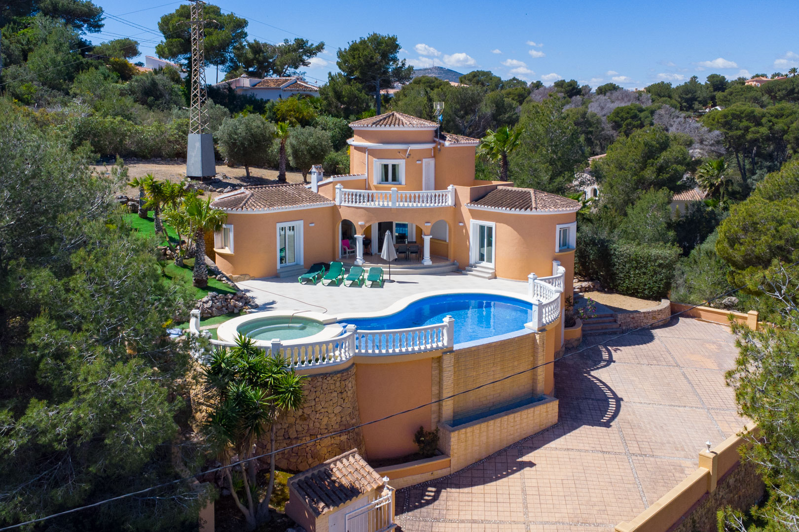 Luchtfoto van de mediterrane villa met een niervormig zwembad, geïntegreerde jacuzzi en witte balustrades, omgeven door pijnbomen en een bestrate oprit.
