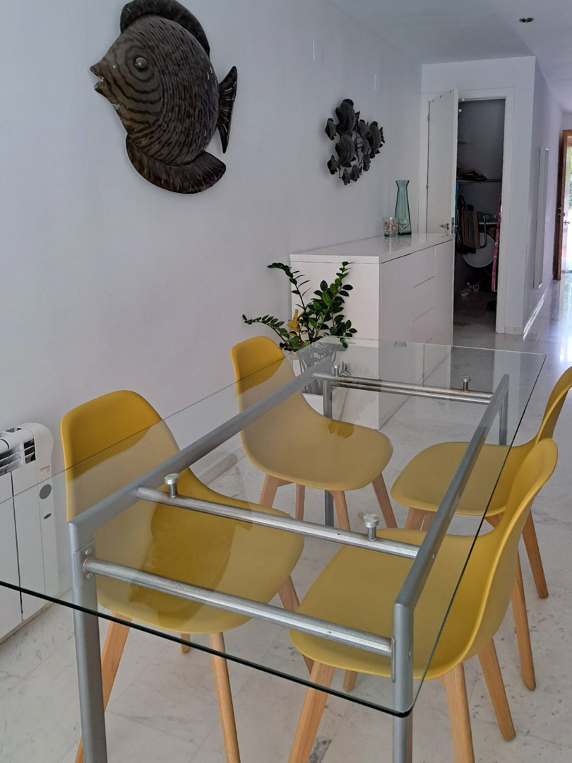 Eethoek met een glazen tafel en gele stoelen op een marmeren vloer. De ruimte is voorzien van een wit dressoir, elektrische radiator en wanddecoratie in zeethema.