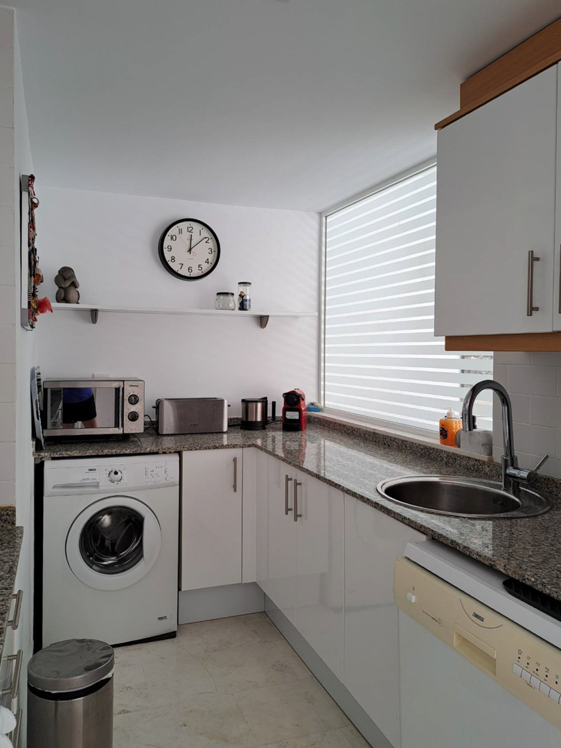 Functionele L-vormige keuken met granieten werkblad, witte kasten, geïntegreerde wasmachine en een groot raam voor natuurlijke lichtinval.
