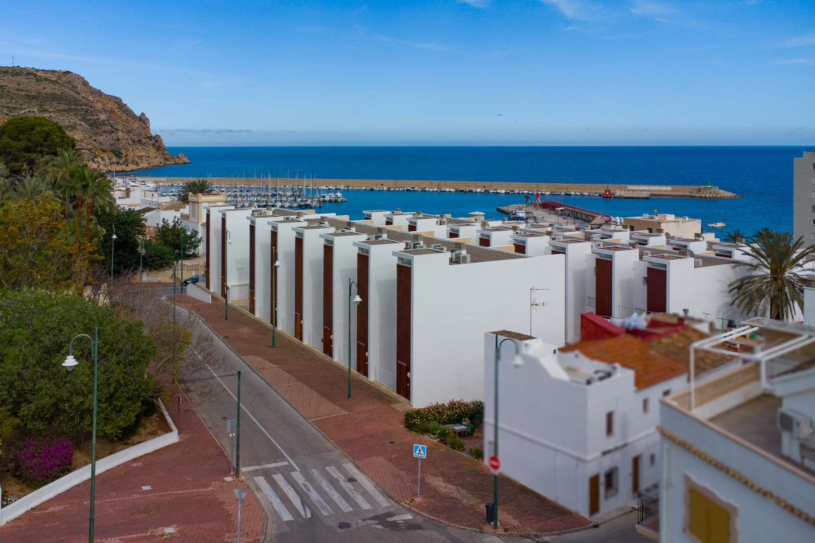 Panoramisch uitzicht op de haven van Jávea en de Middellandse Zee, met moderne witte gebouwen, kustwegen en de nabijgelegen jachthaven in de zon.