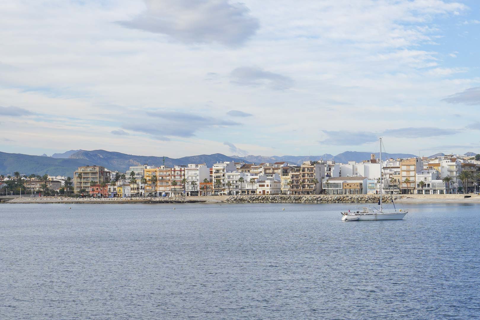 Panoramisch uitzicht op de kustlijn van de haven van Jávea vanaf het water, met de bebouwing aan de boulevard, een zeilboot en de bergen op de achtergrond.