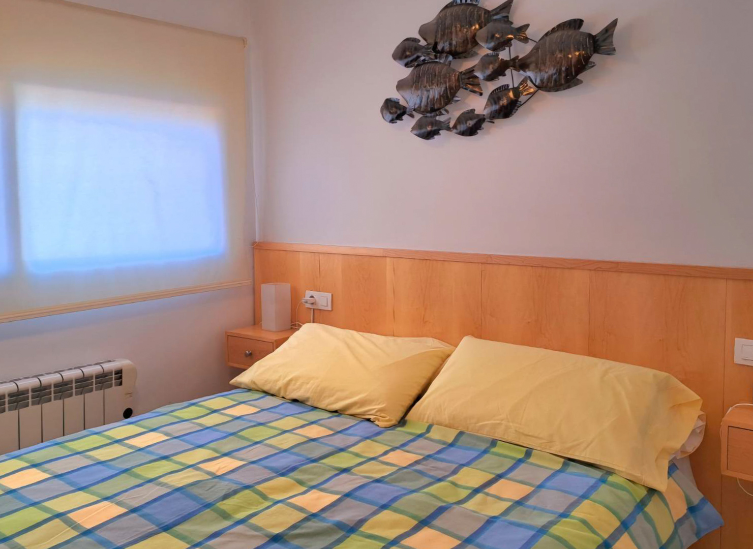 Slaapkamer met geïntegreerd houten hoofdbord, bijpassende nachtkastjes en metalen wanddecoratie. Voorzien van een elektrische radiator en raam met rolgordijn.