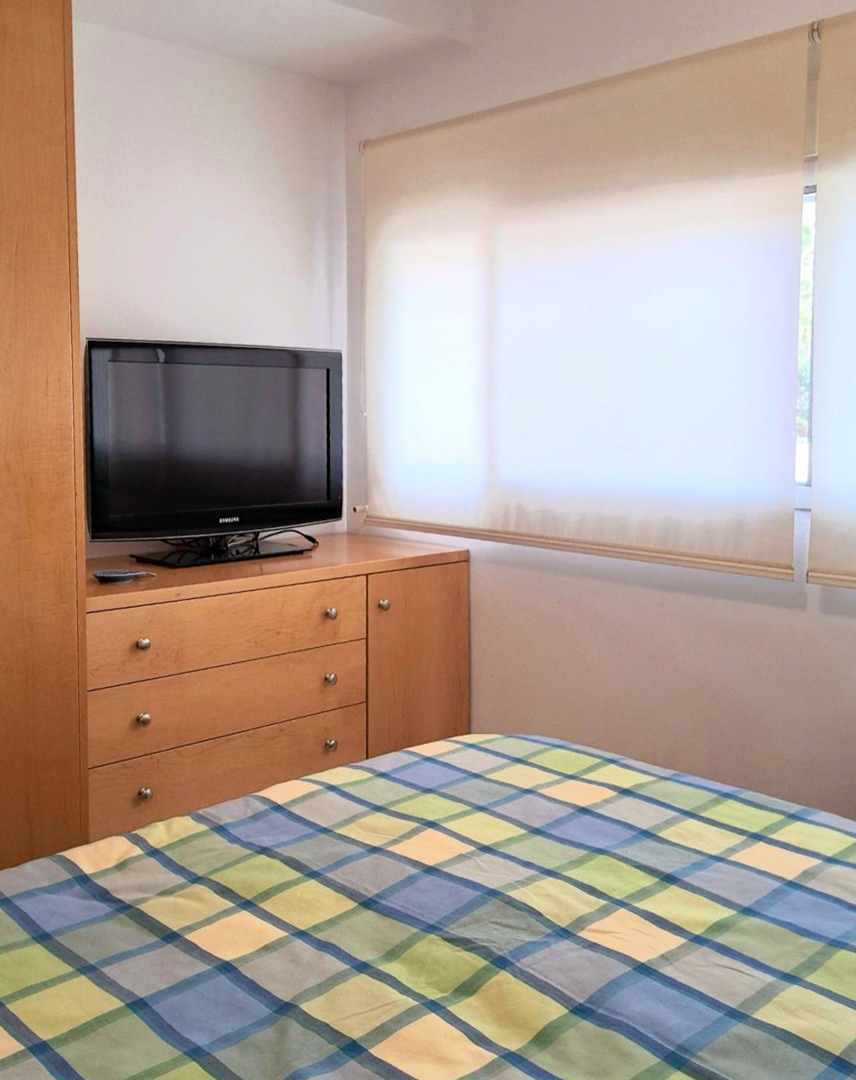 Slaapkamer met op maat gemaakte houten meubels, televisie op een ladekast en raam met lichtdoorlatende rolgordijnen.