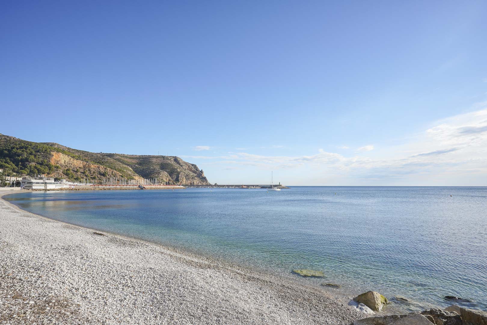 Uitzicht op het kiezelstrand in Jávea, met helder zeewater en de jachthaven gelegen aan de voet van de kliffen van Cabo de San Antonio.