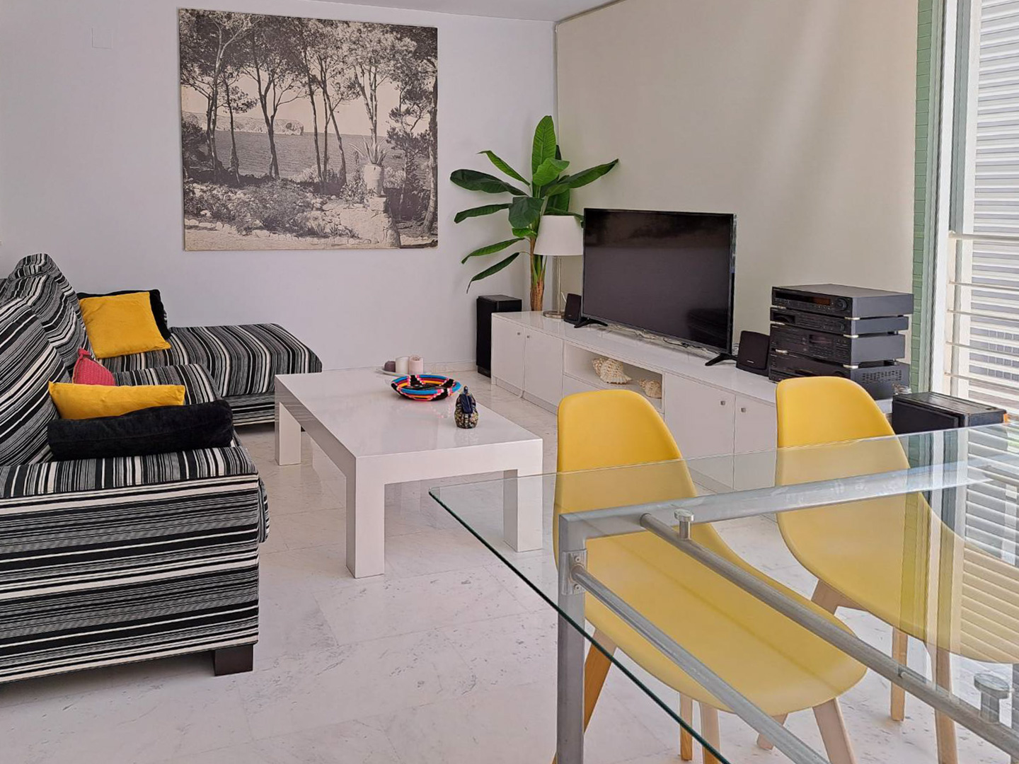 Woon-eetkamer met marmeren vloer, gestreepte bank en glazen eettafel met gele stoelen. De ruimte is voorzien van een wit tv-meubel en geniet van natuurlijk licht.