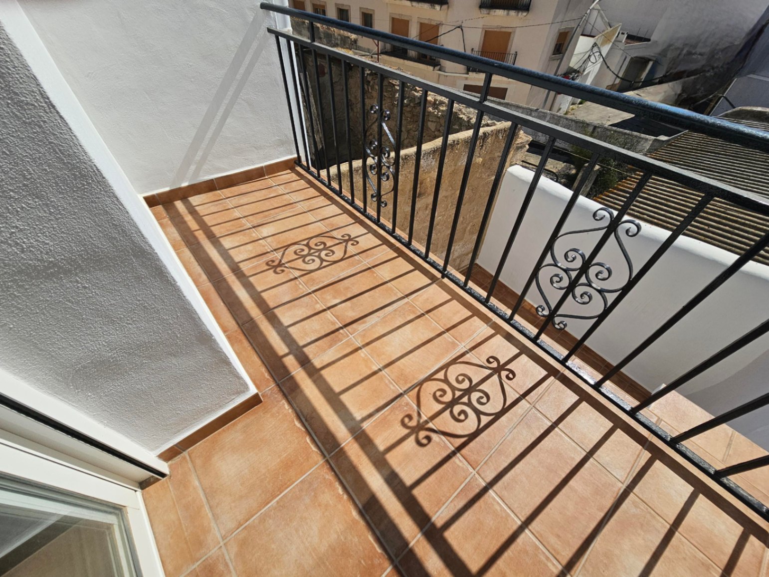 Balkon met terracotta tegelvloer en zwart smeedijzeren hekwerk met decoratieve details, uitkijkend over de traditionele dorpsarchitectuur.