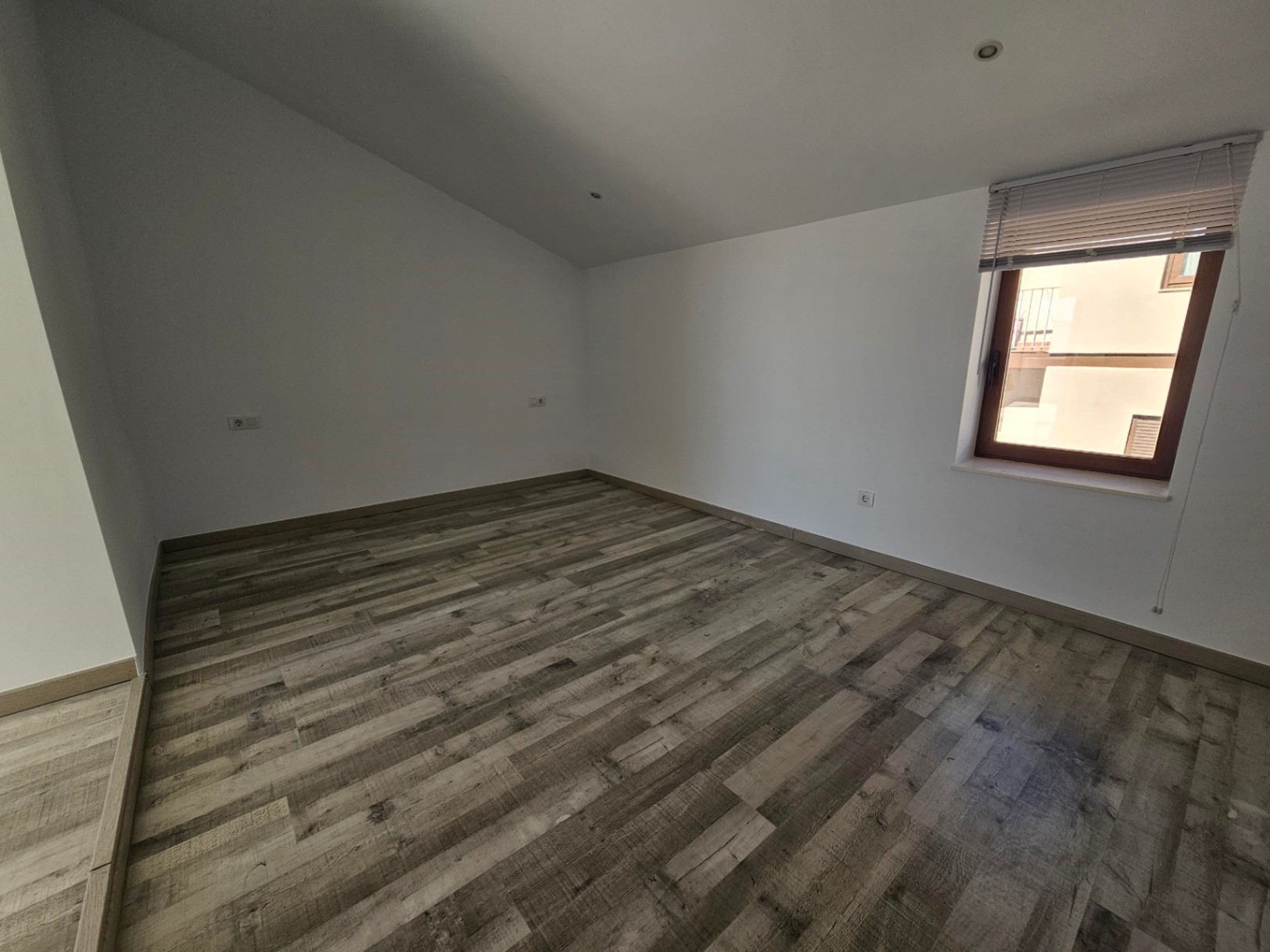 Kamer met schuin plafond, laminaatvloer met houtoptiek en inbouwspots. Voorzien van een houten raamkozijn met jaloezieën voor natuurlijk licht.