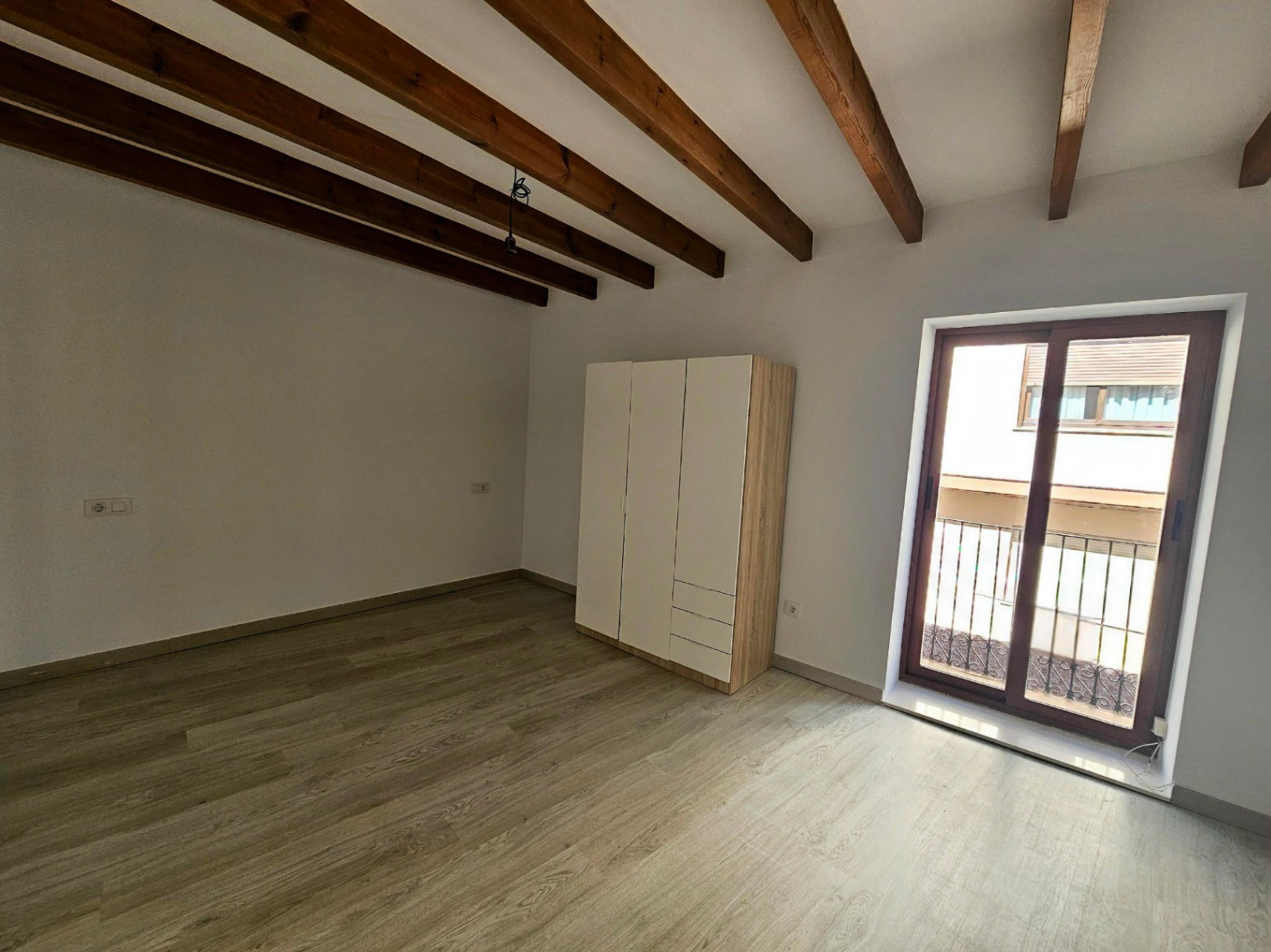 Lichte slaapkamer met authentieke houten plafondbalken, een lichte laminaatvloer en toegang tot een privébalkon via een grote glazen schuifdeur.