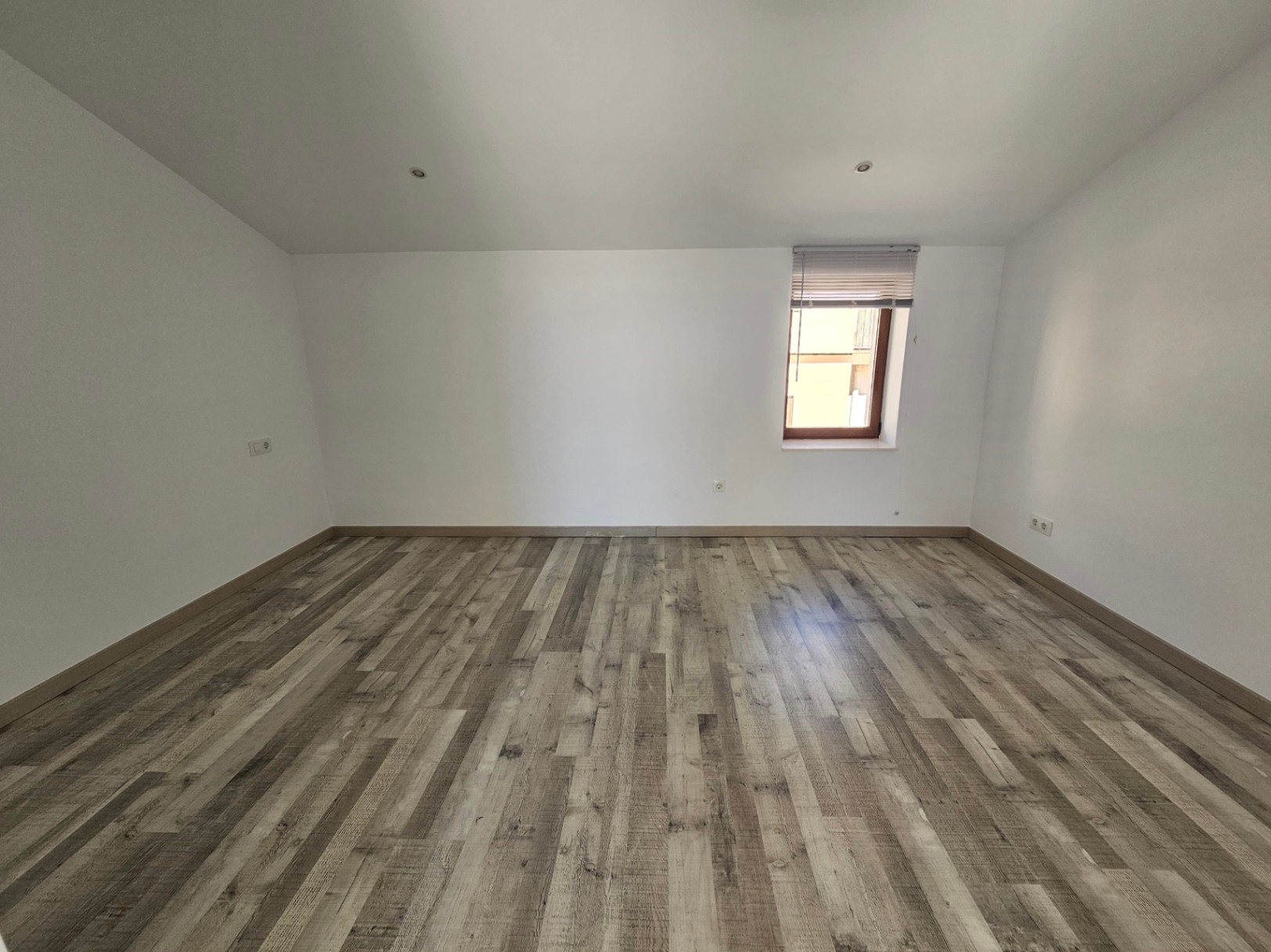Lichte zolderkamer met schuin plafond, grijze laminaatvloer en inbouwspots. Een houten raamkozijn met jaloezie biedt natuurlijk licht in deze gerenoveerde ruimte.