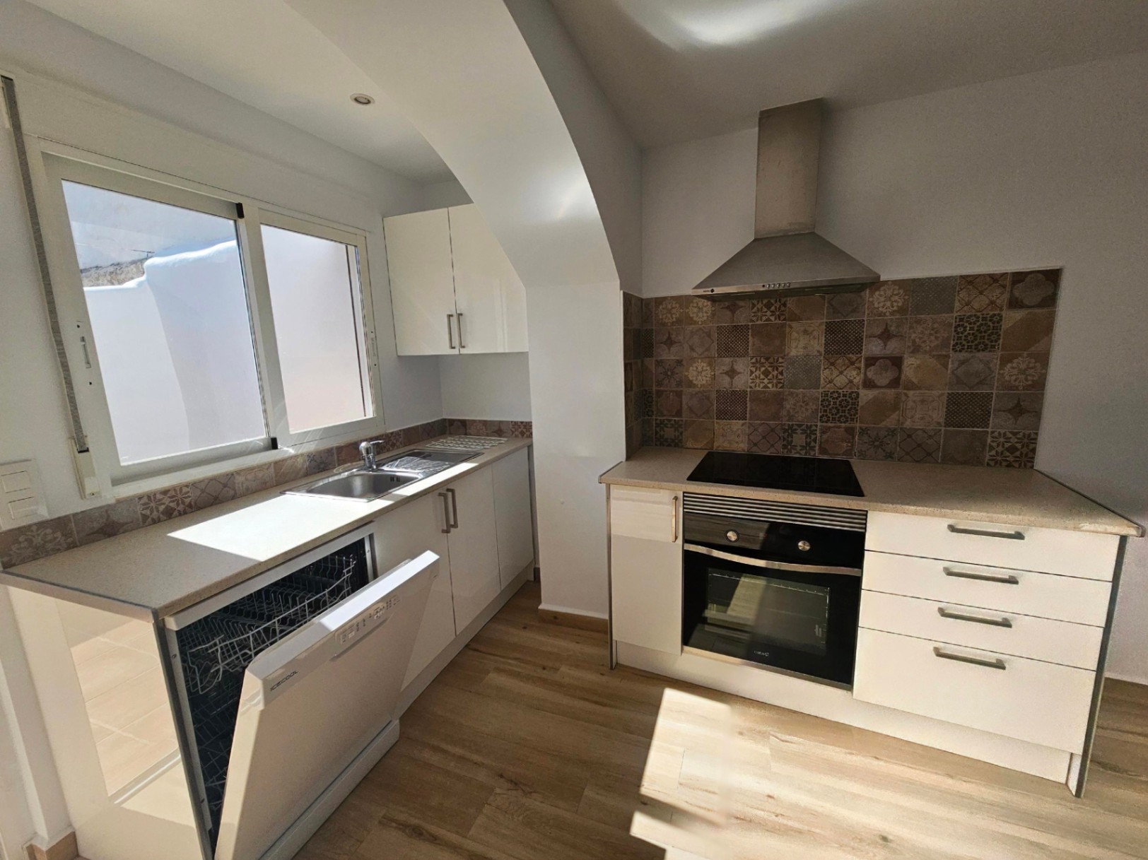 Moderne keuken met witte kasten, aanrechtblad met steeneffect en decoratieve tegelwand. Voorzien van inbouwapparatuur en lichtinval via het raam naar de patio.