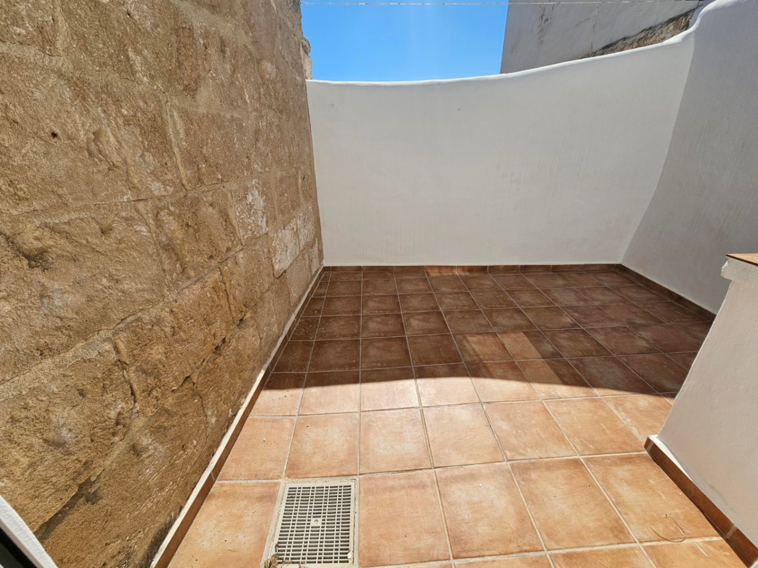 Privé buitenpatio met terracotta vloertegels, een authentieke natuurstenen muur en witte wanden onder een helderblauwe hemel.
