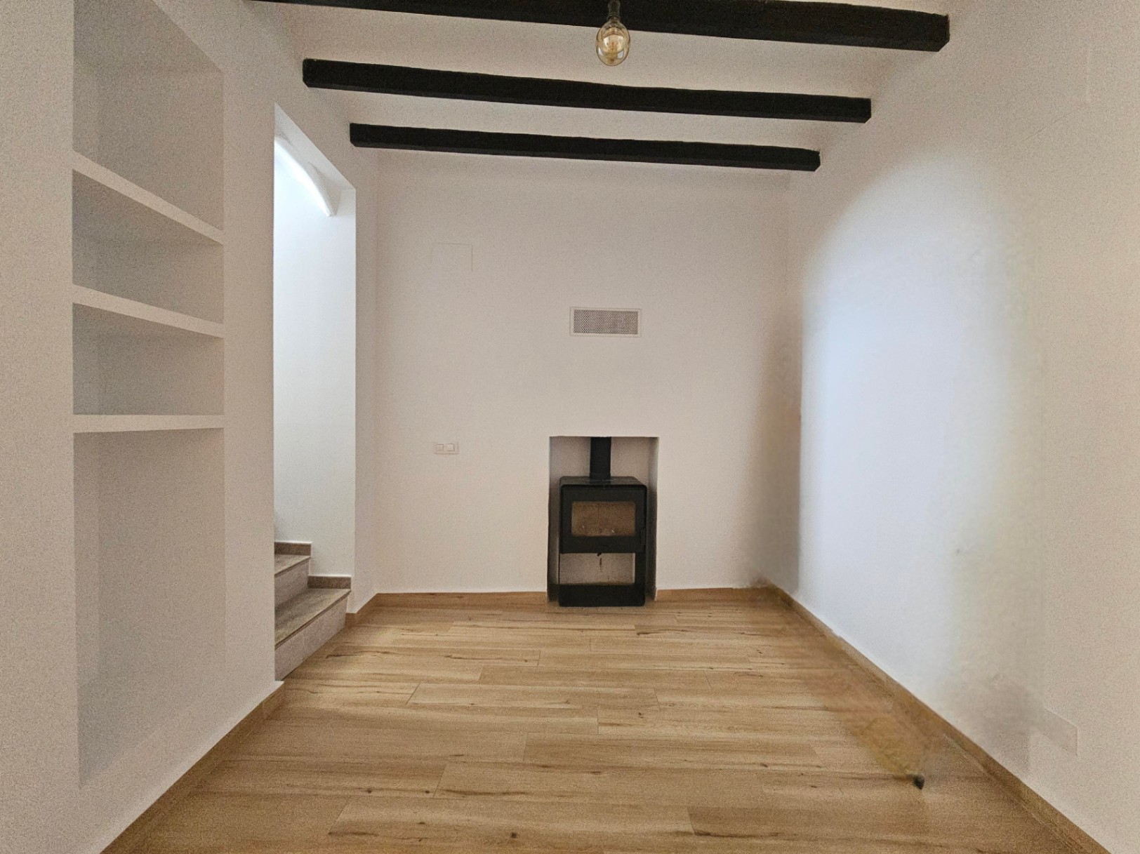 Woonkamer met ingebouwde houtkachel, donkere plafondbalken en lichte houten vloer. Inclusief ingebouwde witte wandkast en toegang tot de trap.