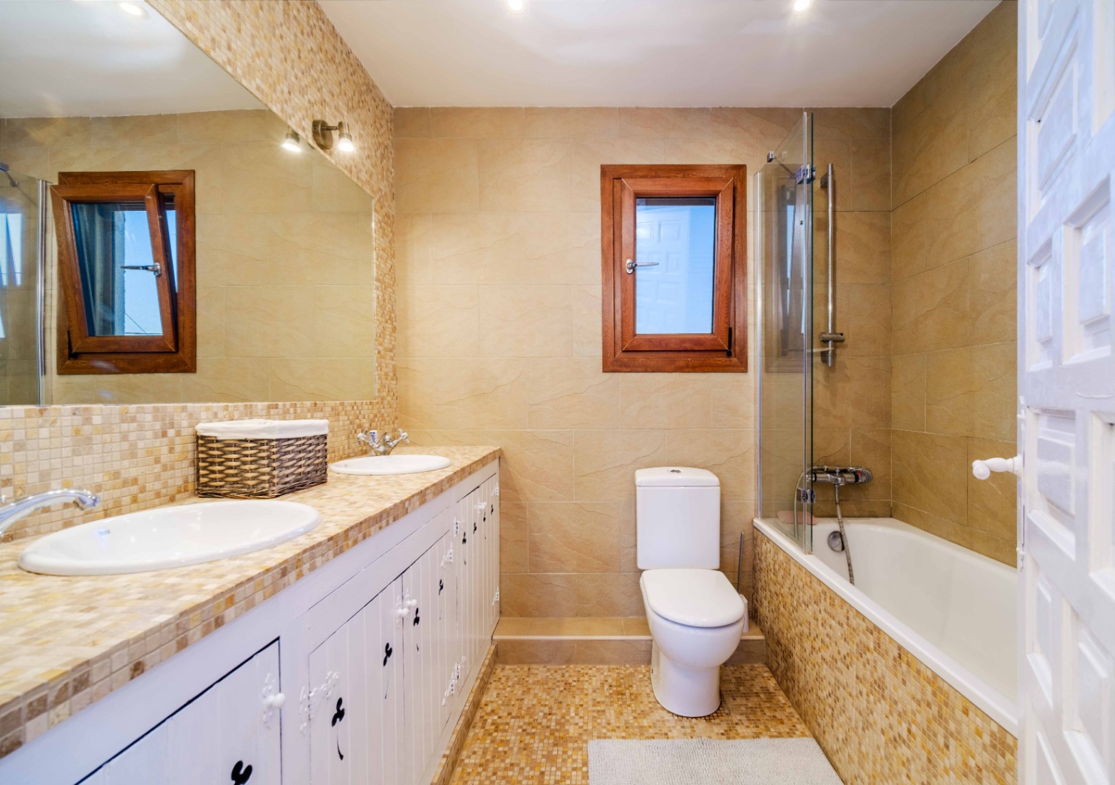 Badkamer met dubbele wastafel, mozaïek aanrechtblad en witte kasten. Voorzien van een ligbad met glazen douchescherm, beige tegels en een raam met houten kozijn.