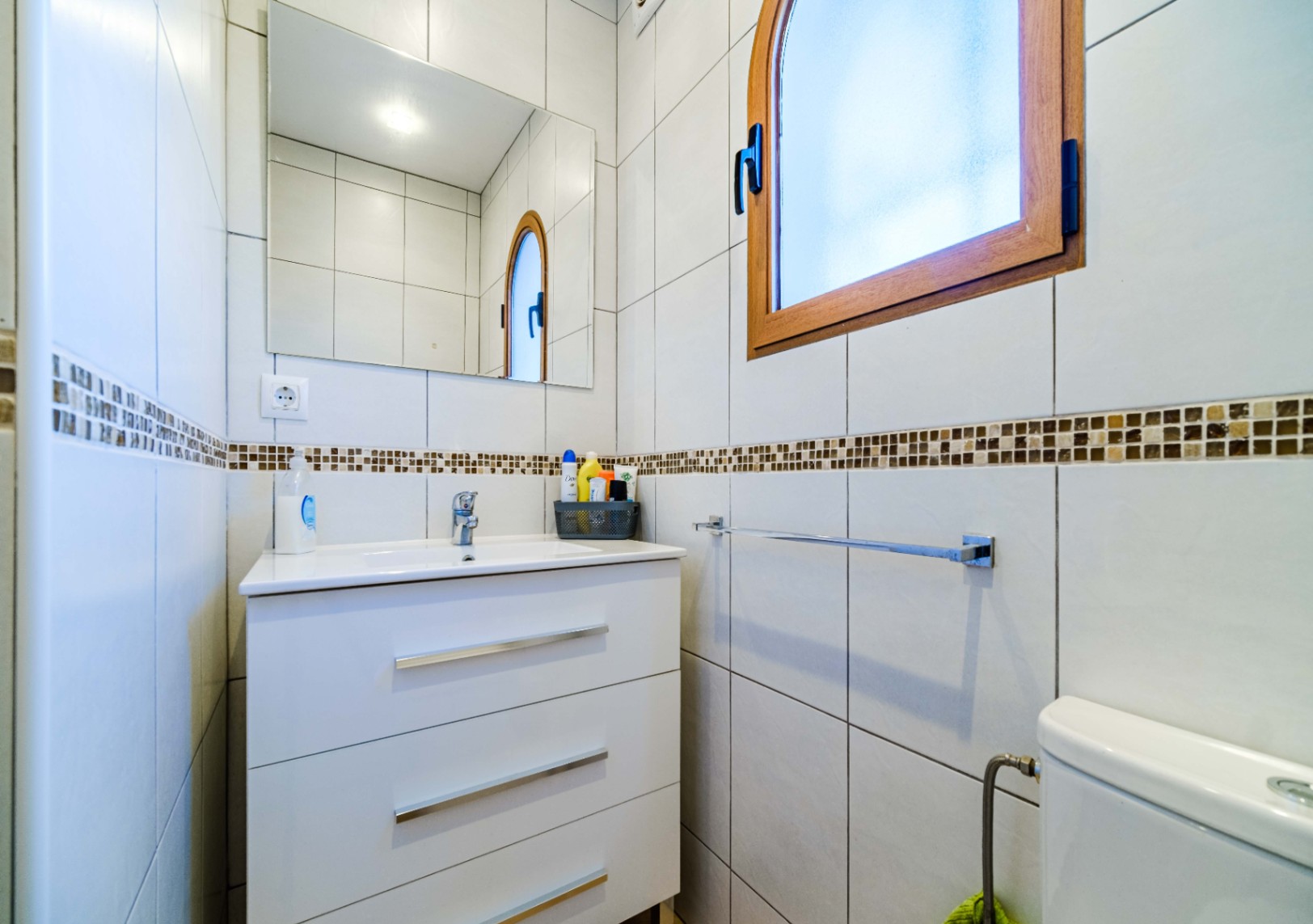 Badkamer met wit wastafelmeubel met drie laden, rechthoekige spiegel en een boogvenster met houten kozijn. Witte wandtegels met een decoratieve mozaïekrand.
