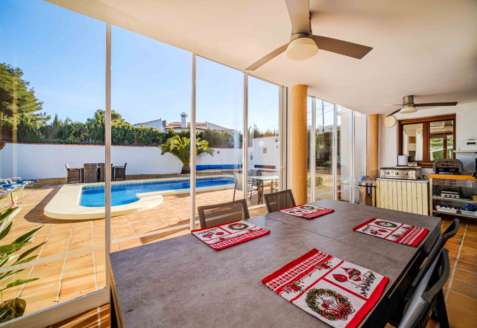 Beglaasd terras met eethoek, plafondventilatoren en buitenkeuken, met direct zicht op het privézwembad en de mediterrane tuin.