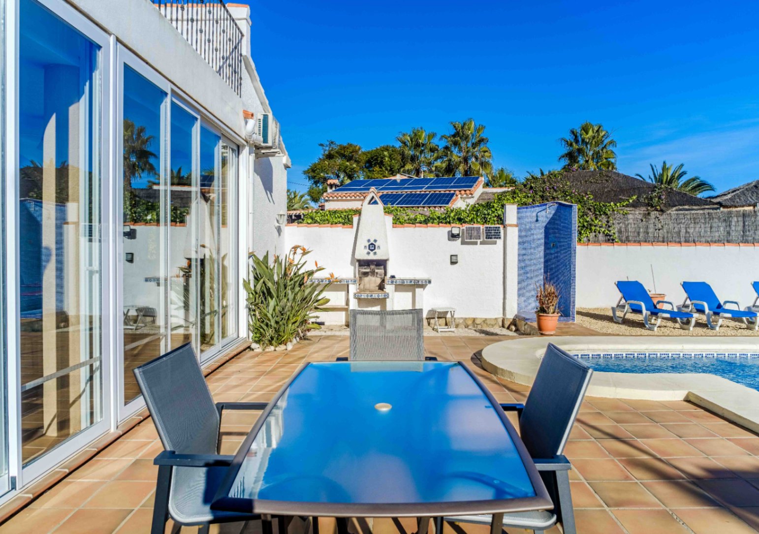 Buitenterras met glazen eettafel, ingebouwde barbecue en blauw betegelde douche bij het zwembad. Voorzien van grote glazen schuifdeuren en zichtbare zonnepanelen.