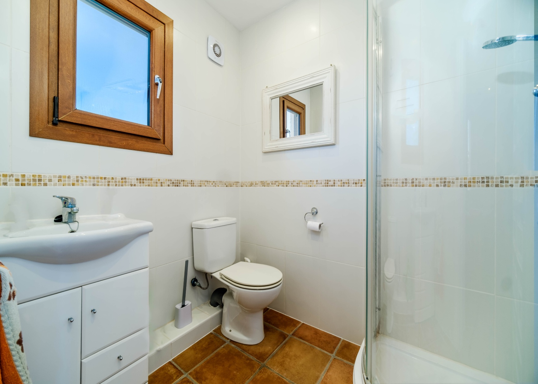 Functionele badkamer met witte tegelwanden, een decoratieve rand, terracotta vloer en een glazen douchecabine.