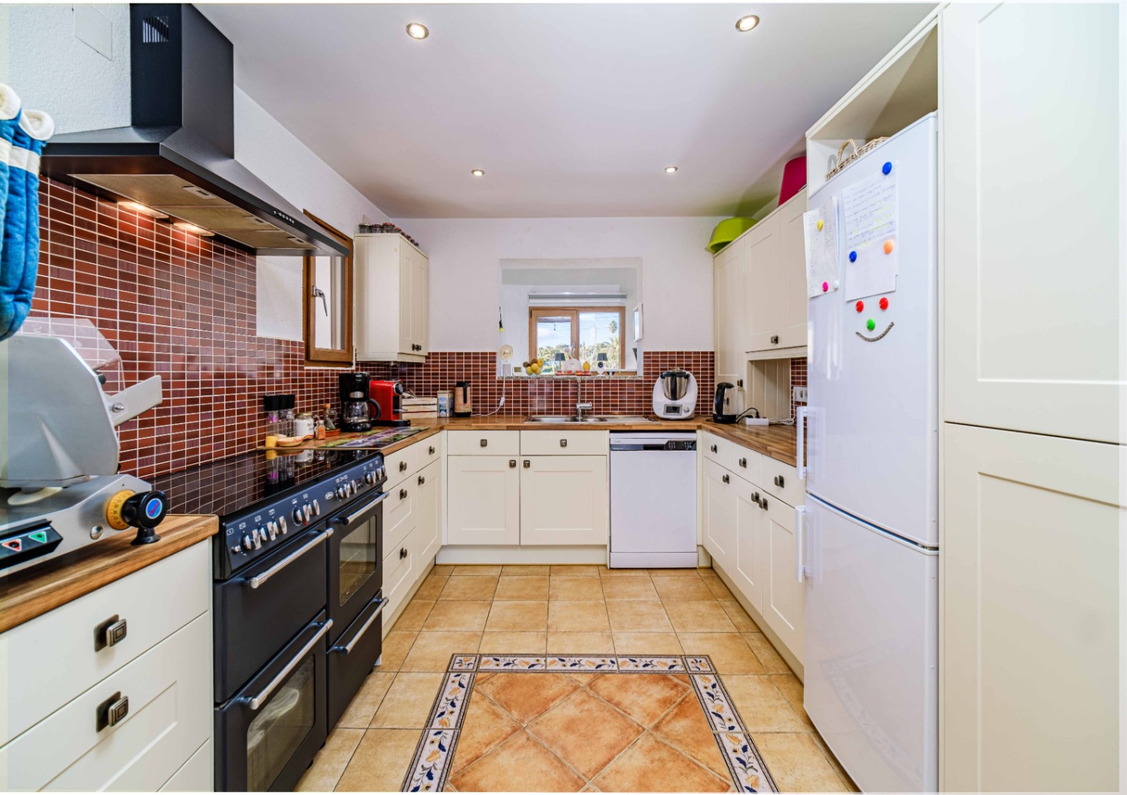 Functionele keuken met witte kasten, houten werkbladen en een bruine tegelwand. Voorzien van een groot zwart fornuis en een terracotta vloer met een decoratieve randafwerking.