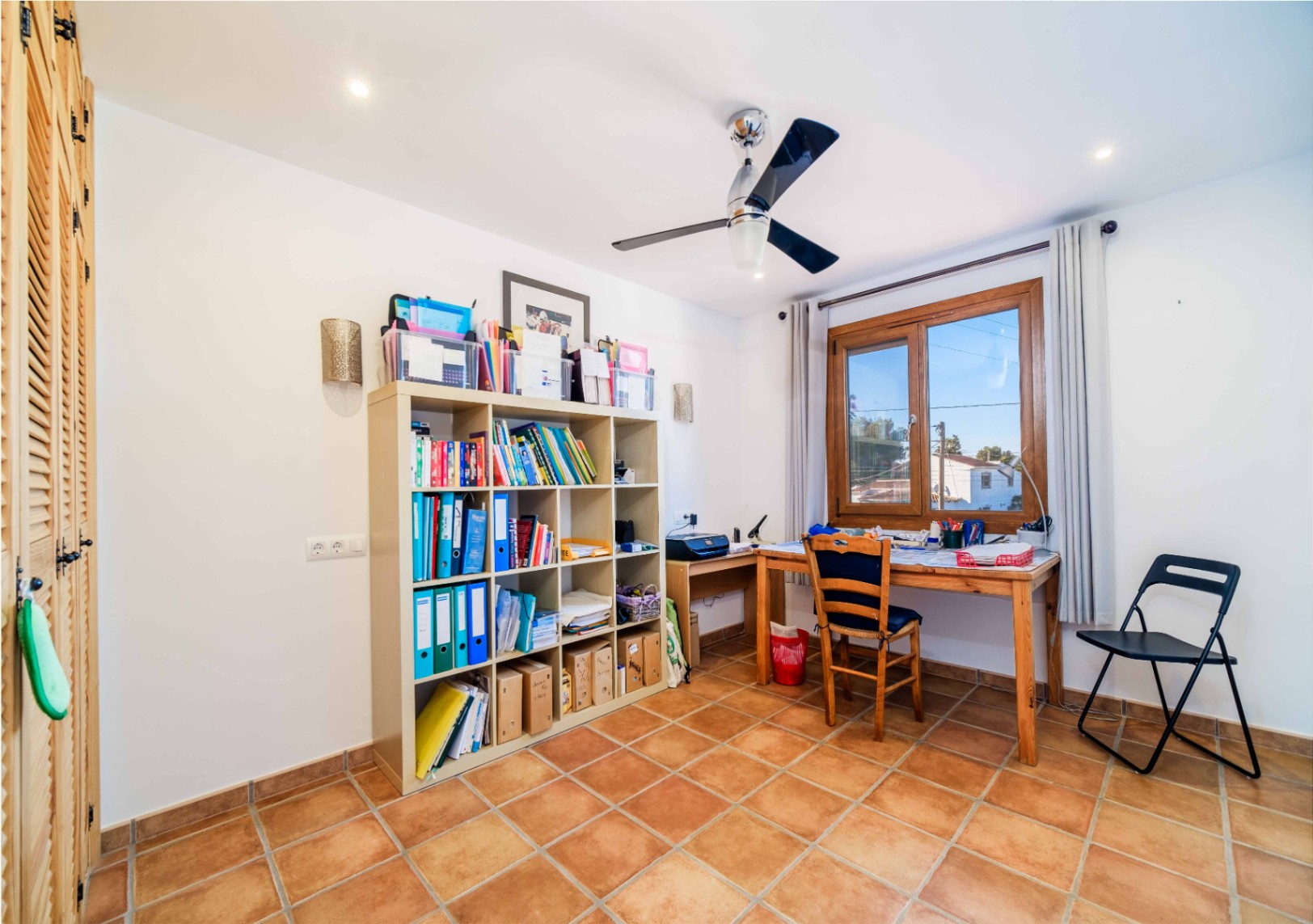Functionele werkkamer met terracotta vloertegels, modulaire kastenwand, houten bureau bij het raam en een moderne plafondventilator.