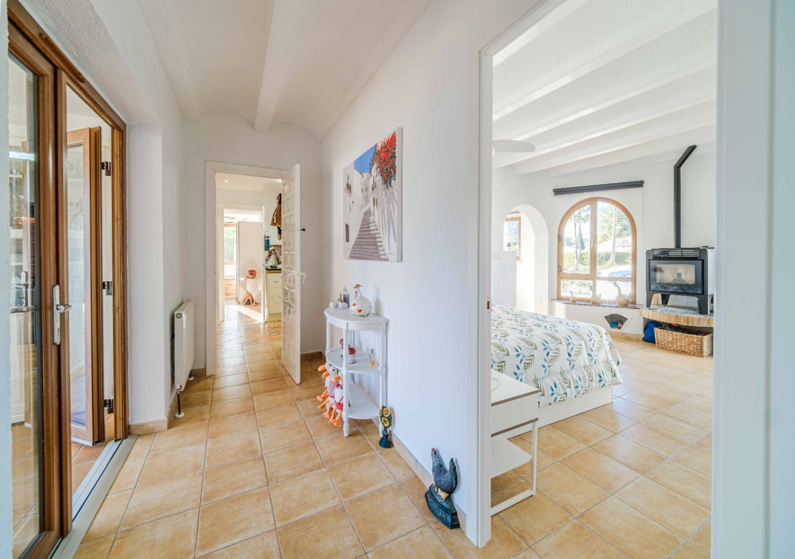 Hal met terracotta vloertegels die leidt naar een lichte slaapkamer met wit balkenplafond, een boogvenster en een vrijstaande houtkachel.