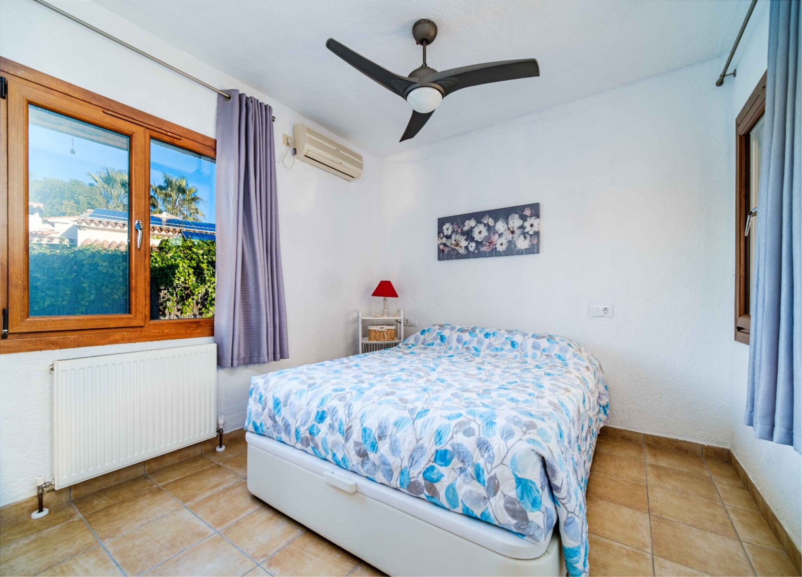 Lichte slaapkamer met tegelvloer, plafondventilator, airconditioning en houten kozijnen met uitzicht op de mediterrane tuin.