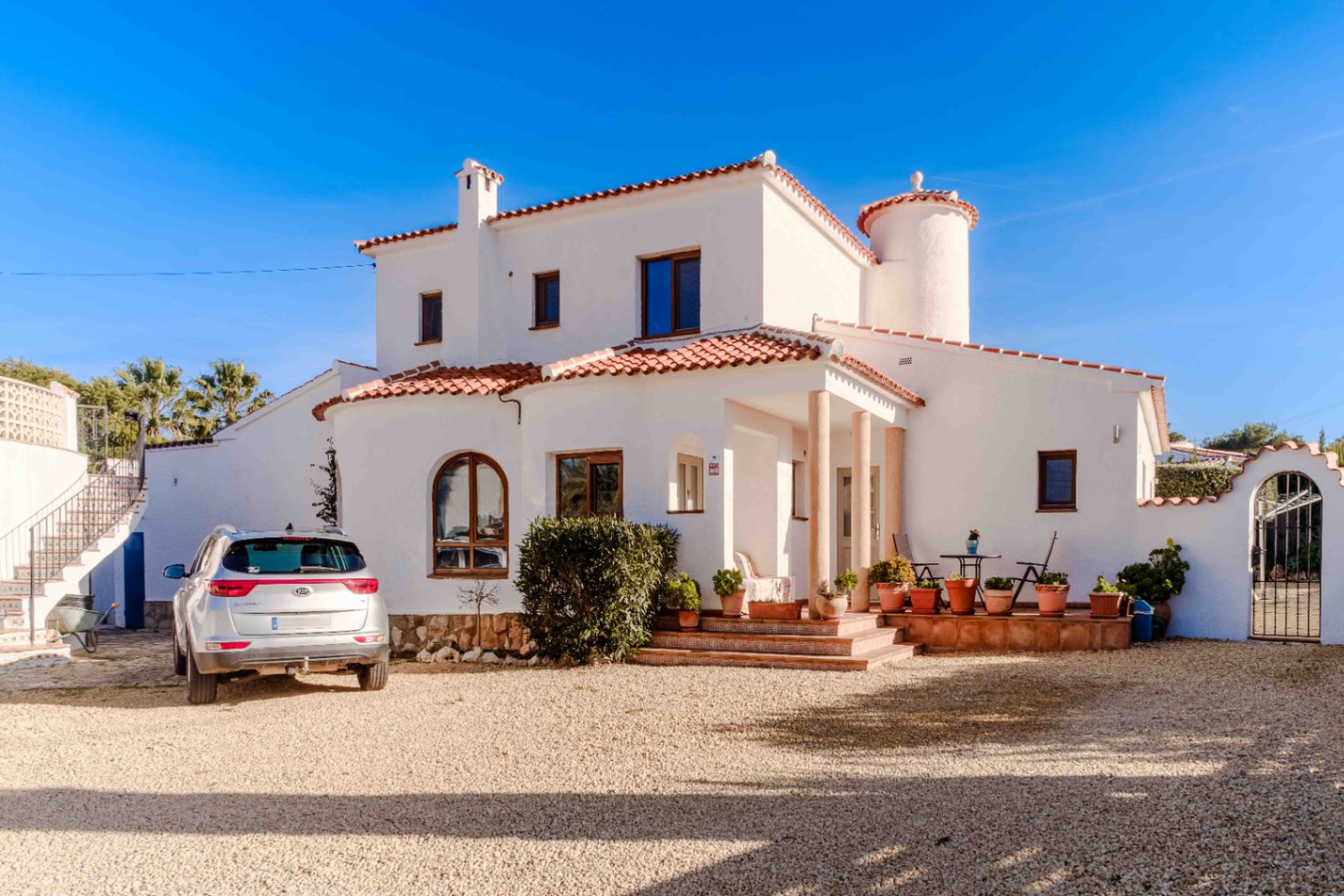 Voorgevel van de mediterrane villa met witte muren, een ronde toren, terracotta dakpannen en een overdekt terras met zuilen aan de oprit van grind.