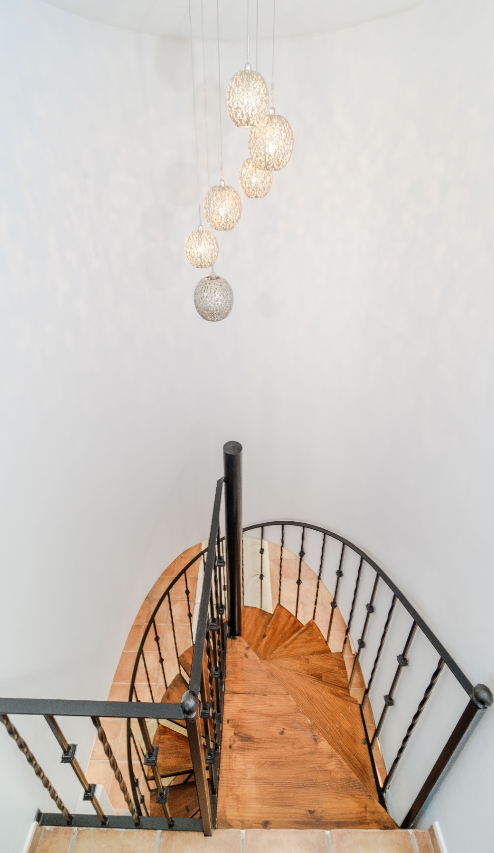 Wenteltrap met massief houten treden en een zwart smeedijzeren leuning, verlicht door een cluster van decoratieve bolvormige hanglampen.