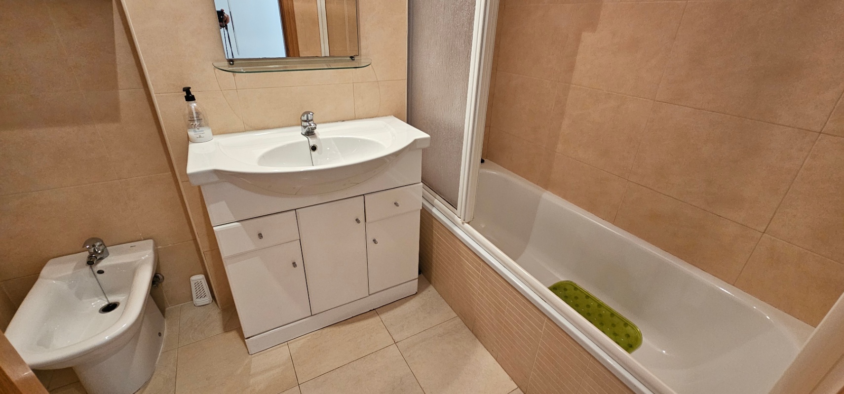 Badkamer met crèmekleurige tegels, voorzien van een ligbad met douchescherm, een bidet en een wit wastafelmeubel met opbergruimte.