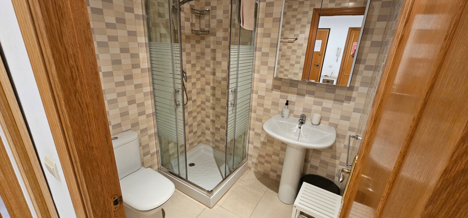Functionele badkamer met een glazen hoekdouche, kolomwastafel en wandtegels in mozaïekstijl in aardetinten.