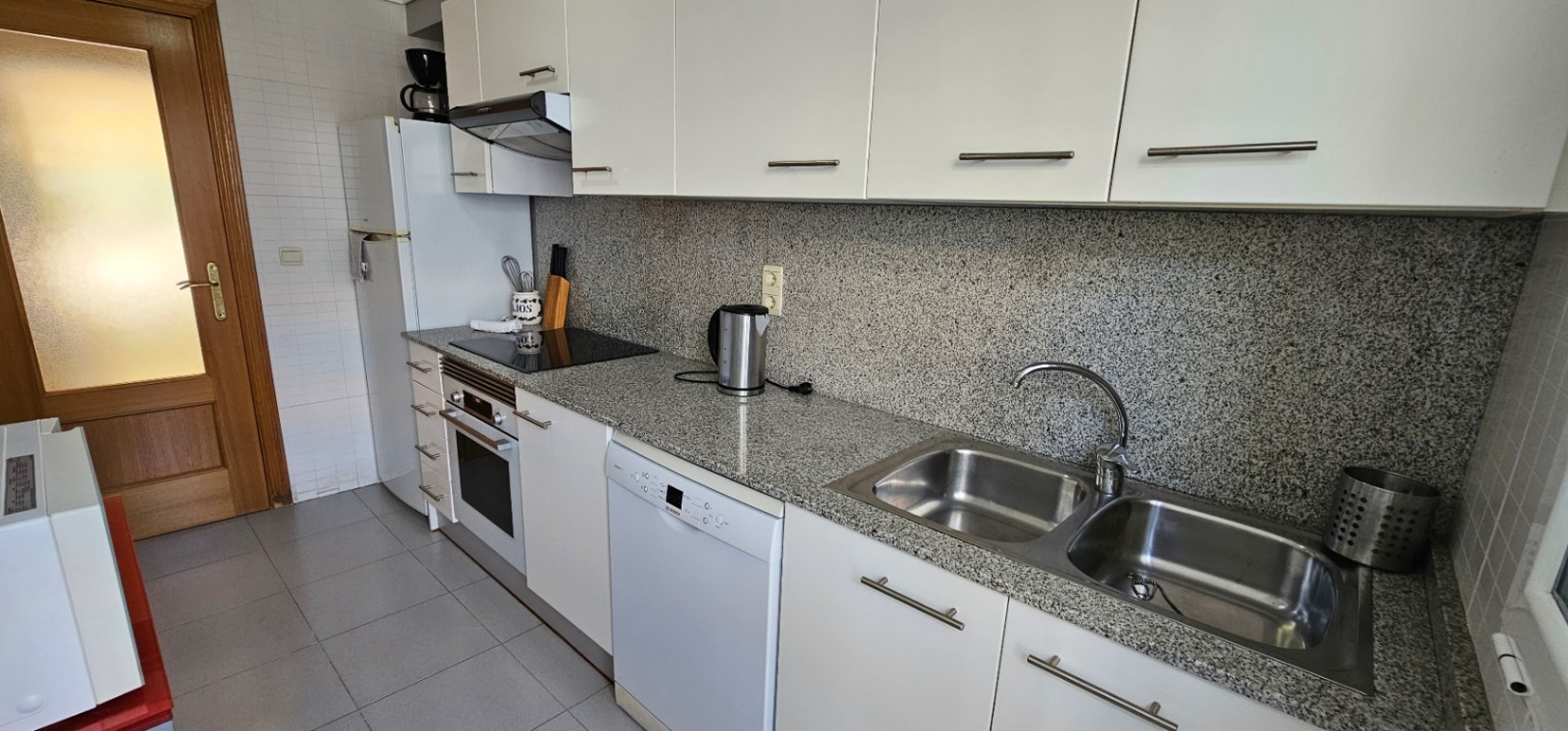 Functionele keuken met grijs granieten aanrechtblad en achterwand, witte kasten, inbouwapparatuur en een dubbele roestvrijstalen spoelbak.