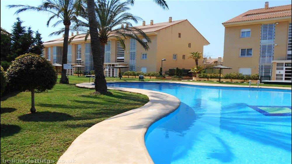 Groot gemeenschappelijk zwembad met organische vormen, omgeven door verzorgde gazons, volwassen palmbomen en mediterrane appartementengebouwen.