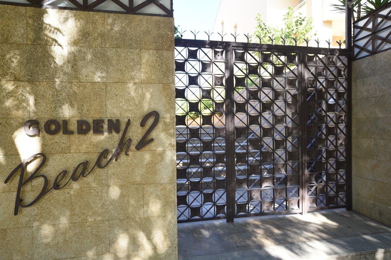 Hoofdingang van het Golden Beach 2 complex, met een natuurstenen muur, metalen belettering en een decoratieve smeedijzeren poort met geometrisch ontwerp.