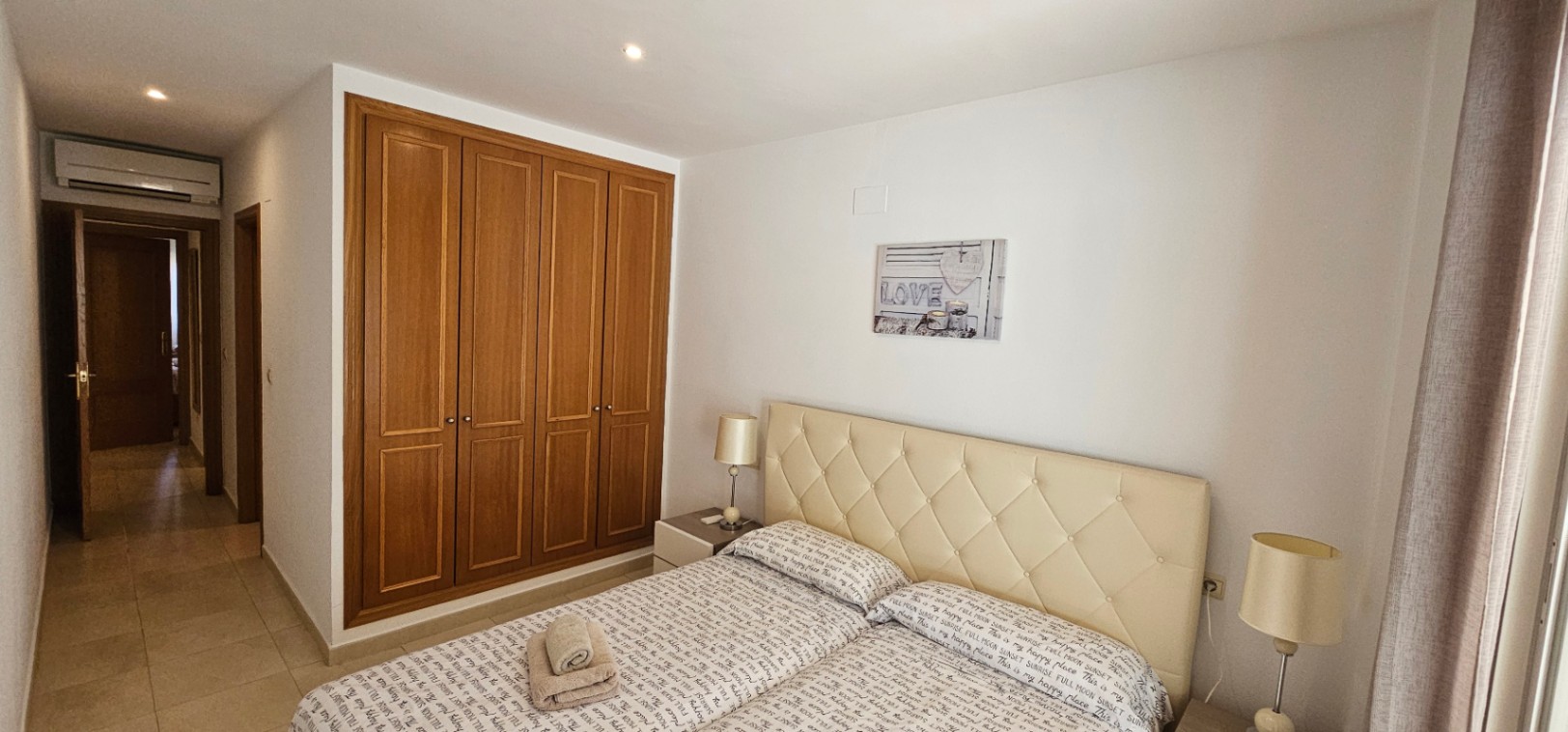 Hoofdslaapkamer met ingebouwde houten kasten, een gecapitonneerd crème hoofdbord en airconditioning boven de toegangsdeur.