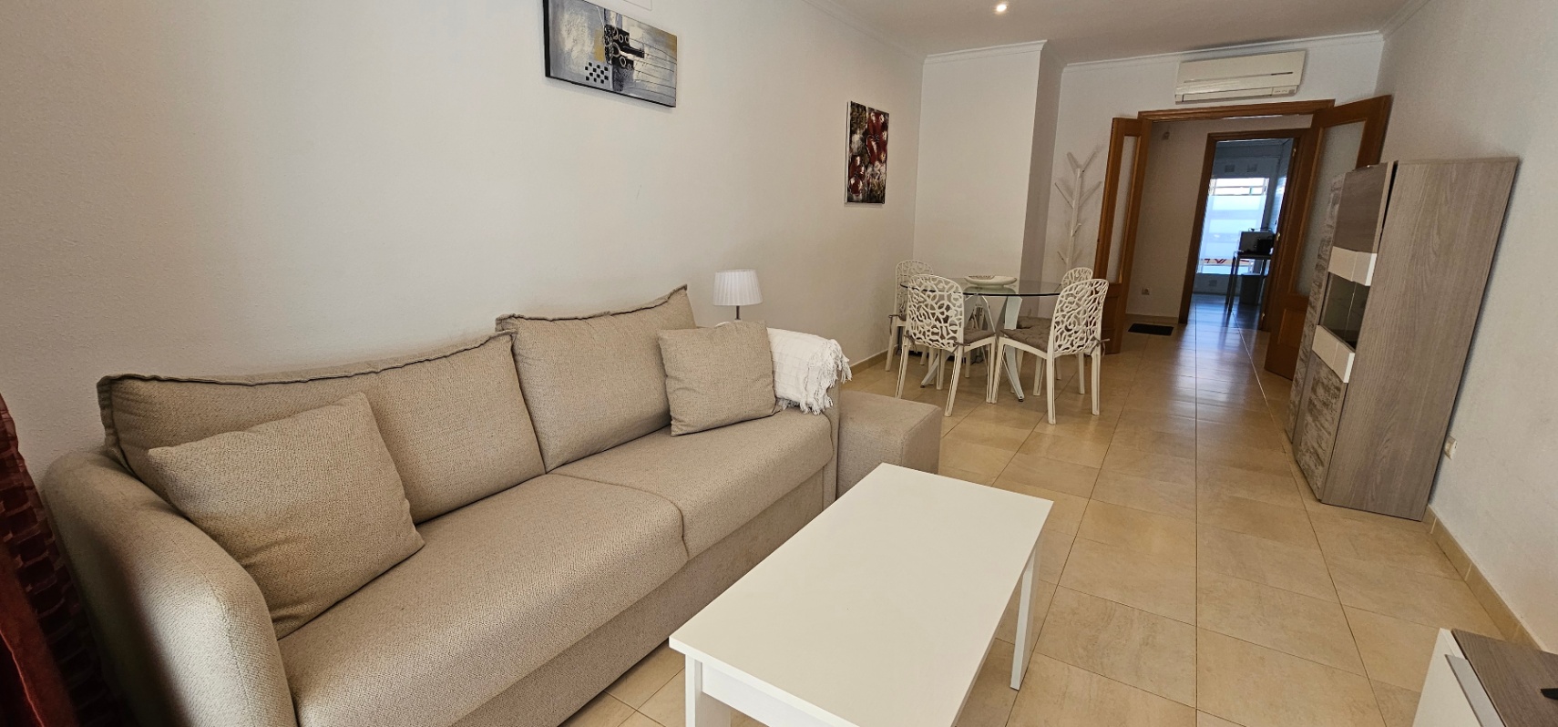Lichte woon- en eetkamer met beige bank, witte salontafel en glazen eettafel. Voorzien van airconditioning en een lichte keramische tegelvloer.