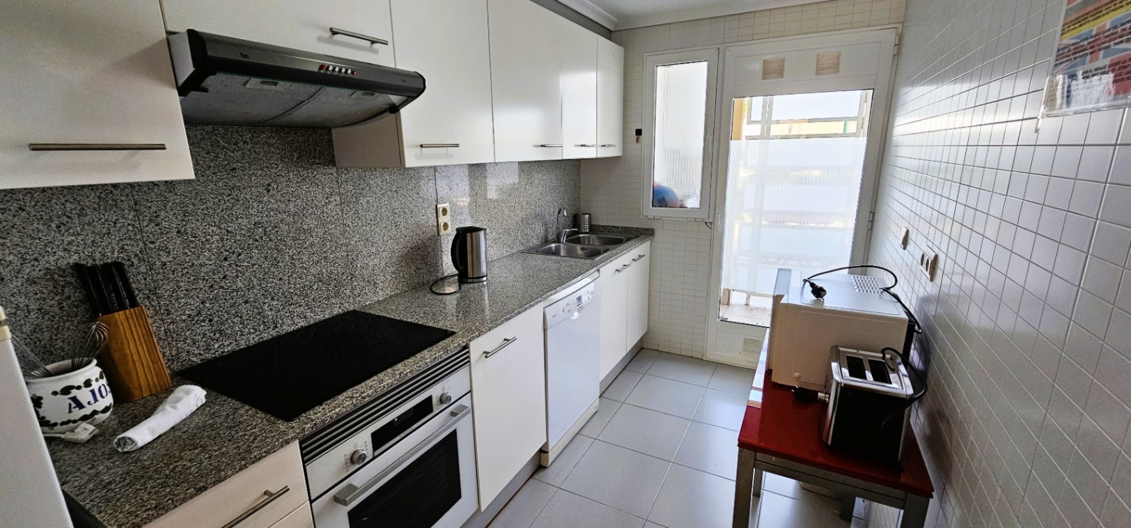 Onafhankelijke keuken met granieten werkblad, witte kasten en inbouwapparatuur. Een glazen deur biedt toegang tot een lichte wasruimte.