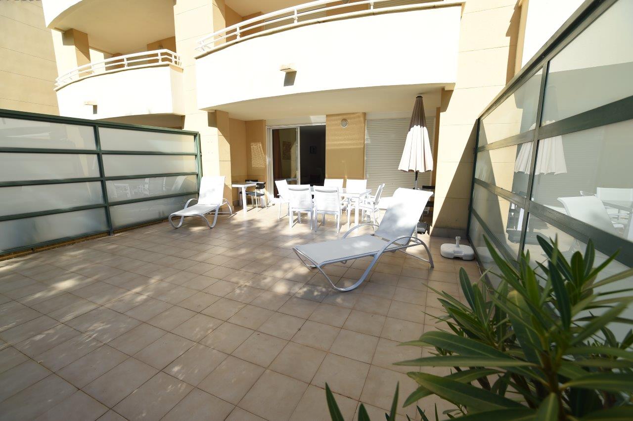 Privé-terras op de begane grond met tegelvloer, witte ligbedden en een eethoek buiten onder de overkapping van het gebouw.